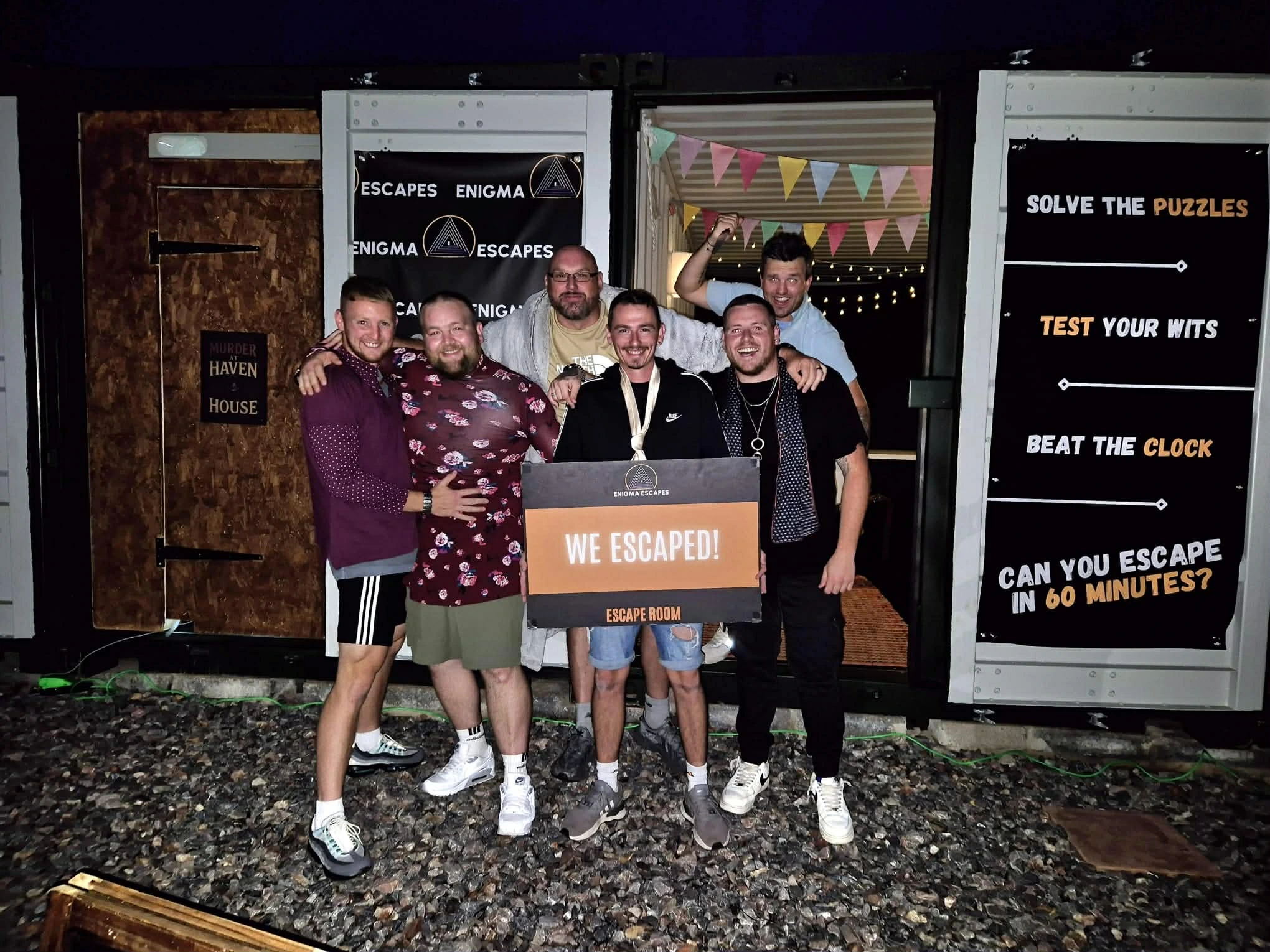 Group of six men smiling and posing outside an escape room holding a sign that says 'WE ESCAPED!'