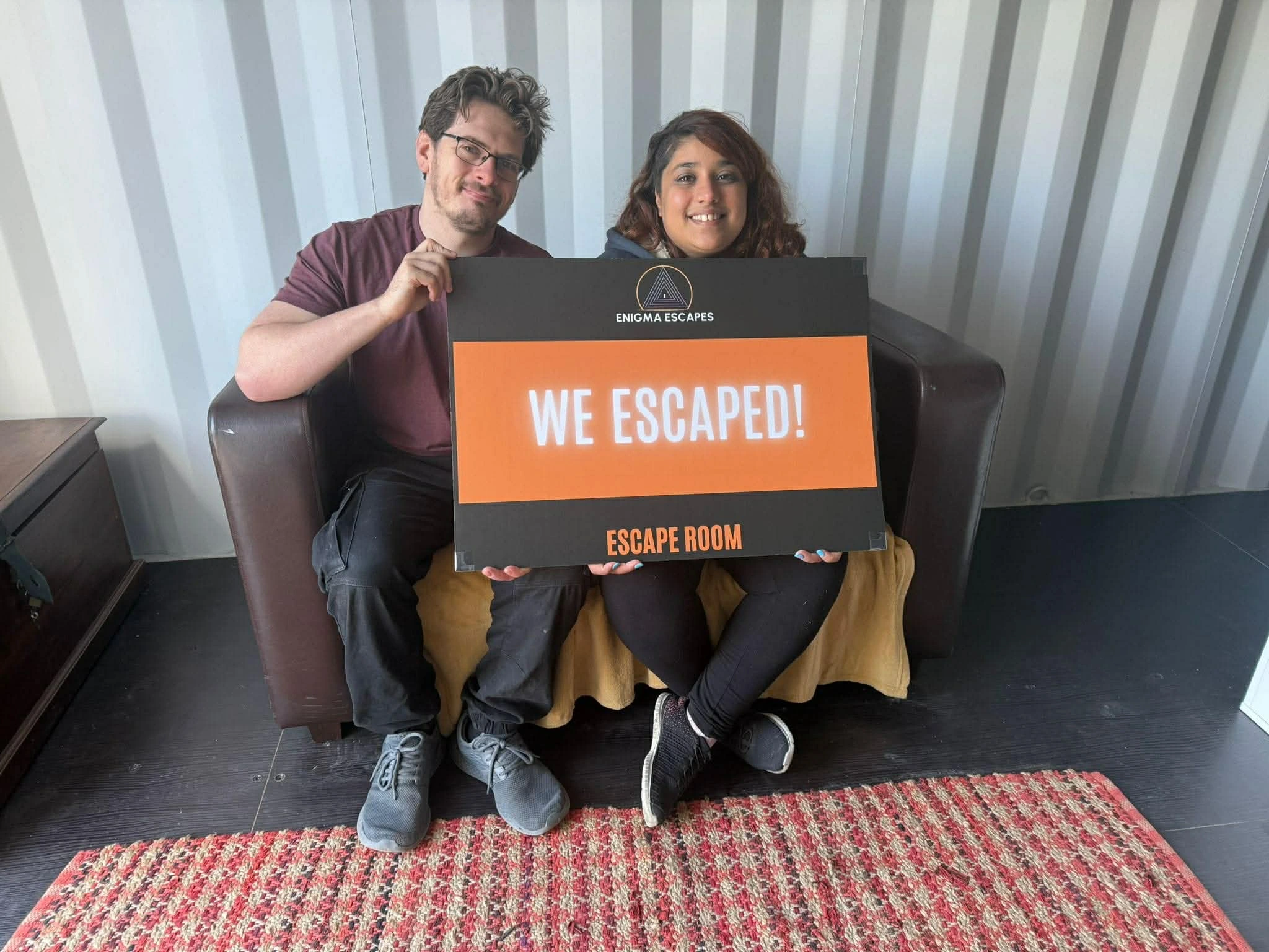 Smiling man and woman sitting on a dark brown couch holding a sign that reads 'WE ESCAPED! ESCAPE ROOM' inside a room with a patterned red and beige rug.