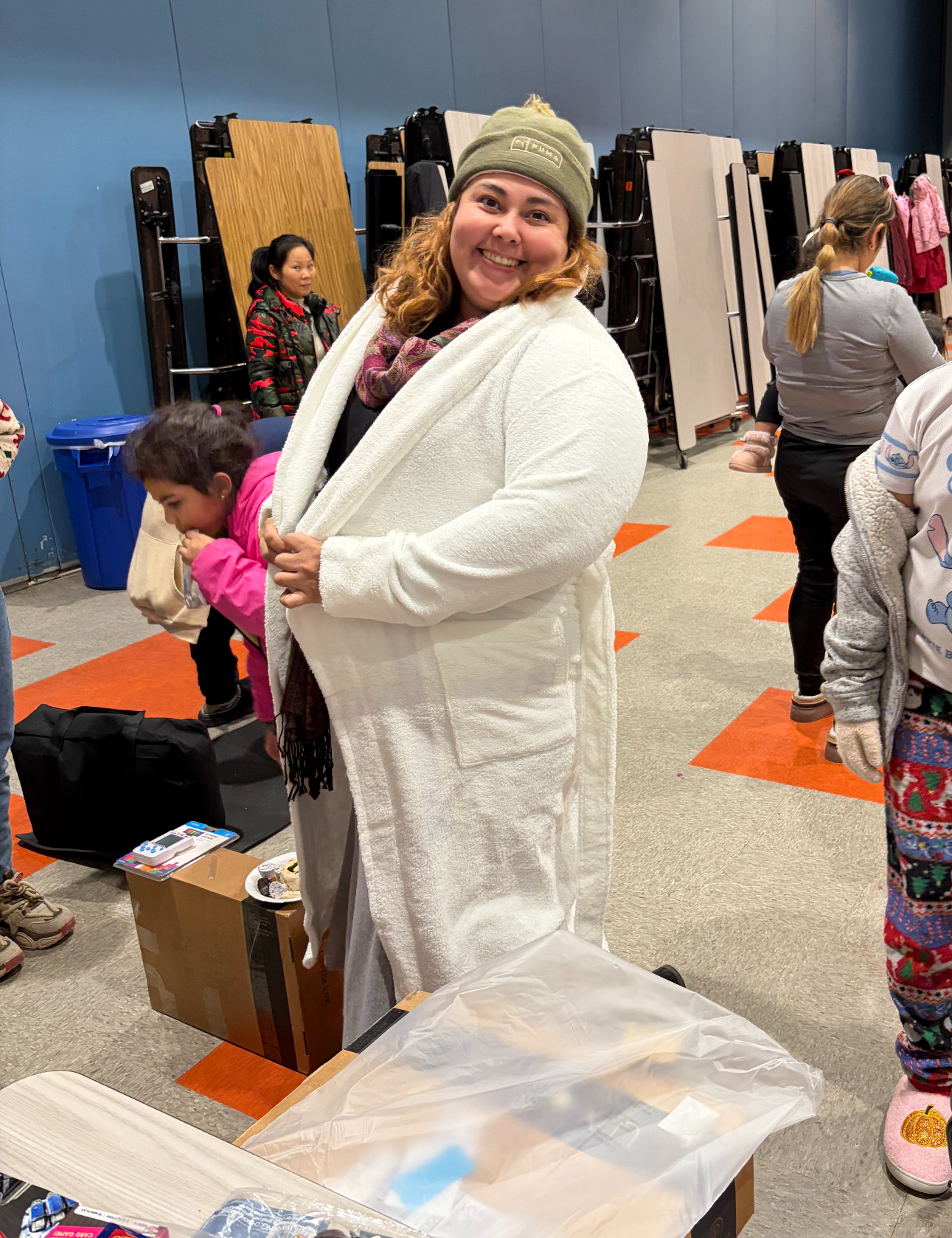 A nonprofit recipient tries on a Luxome robe at a community donation event, smiling as families browse donated goods in the background.