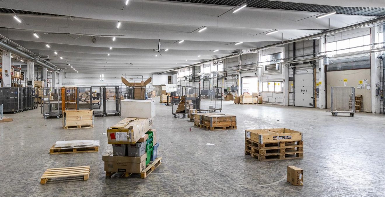 Spacious warehouse interior with scattered wooden pallets, boxes, metal cages, and bright overhead LED lighting.