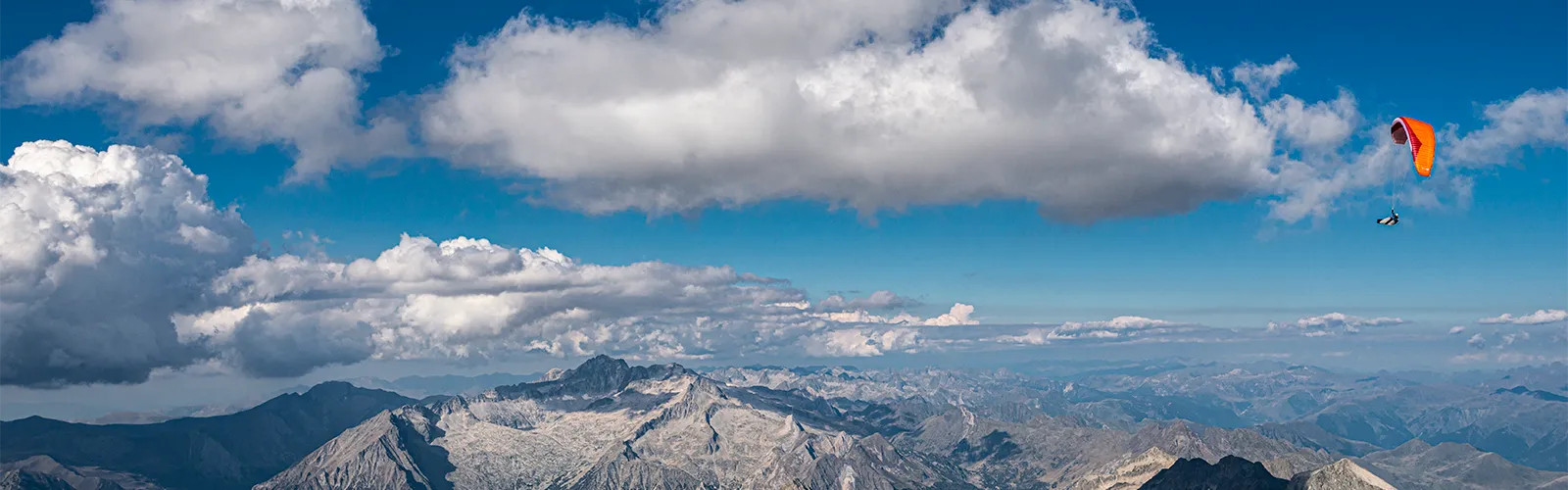 Parapente à voile orange survolant des sommets montagneux rocheux sous un ciel bleu partiellement nuageux.