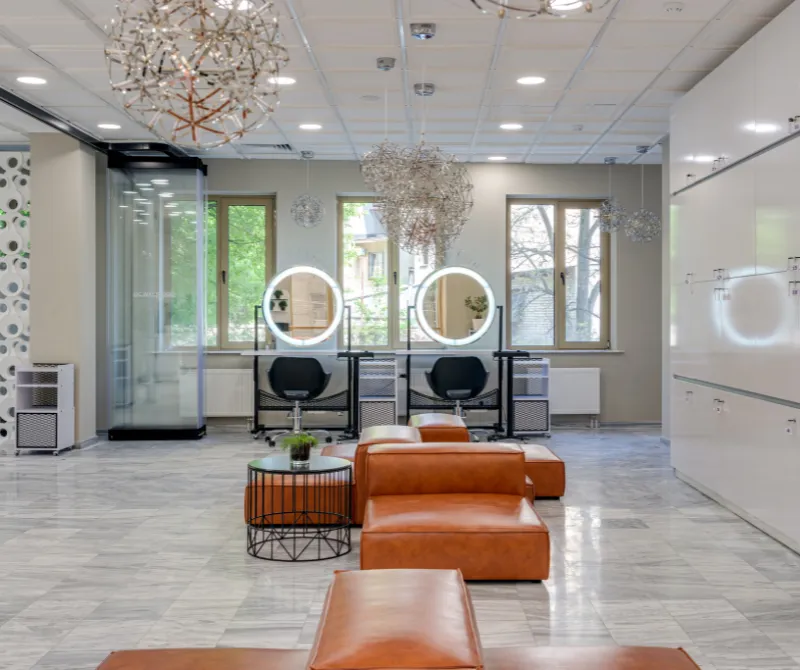 Modern salon interior with round illuminated vanity mirrors, black chairs, leather seating, and large windows.