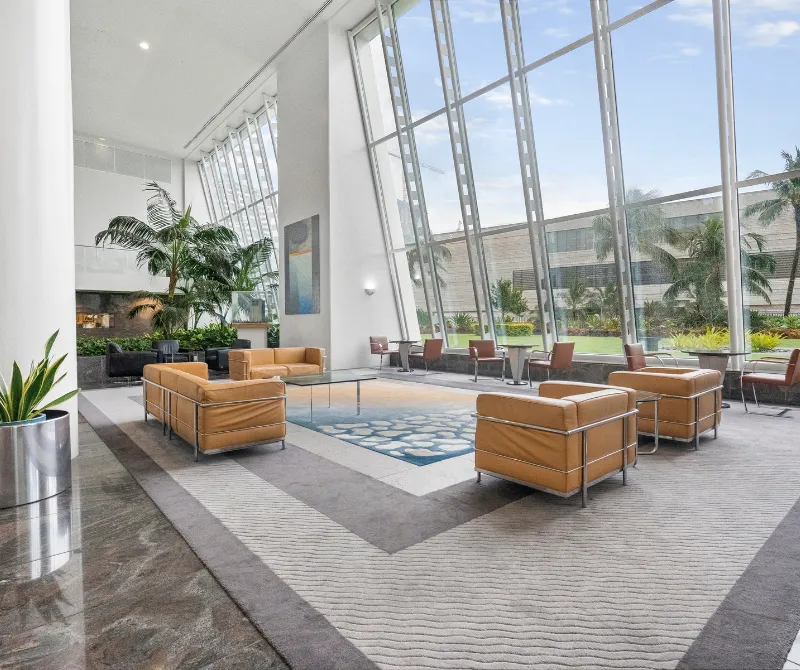 Modern lounge area with tan leather sofas and armchairs, large slanted floor-to-ceiling windows, and indoor plants.