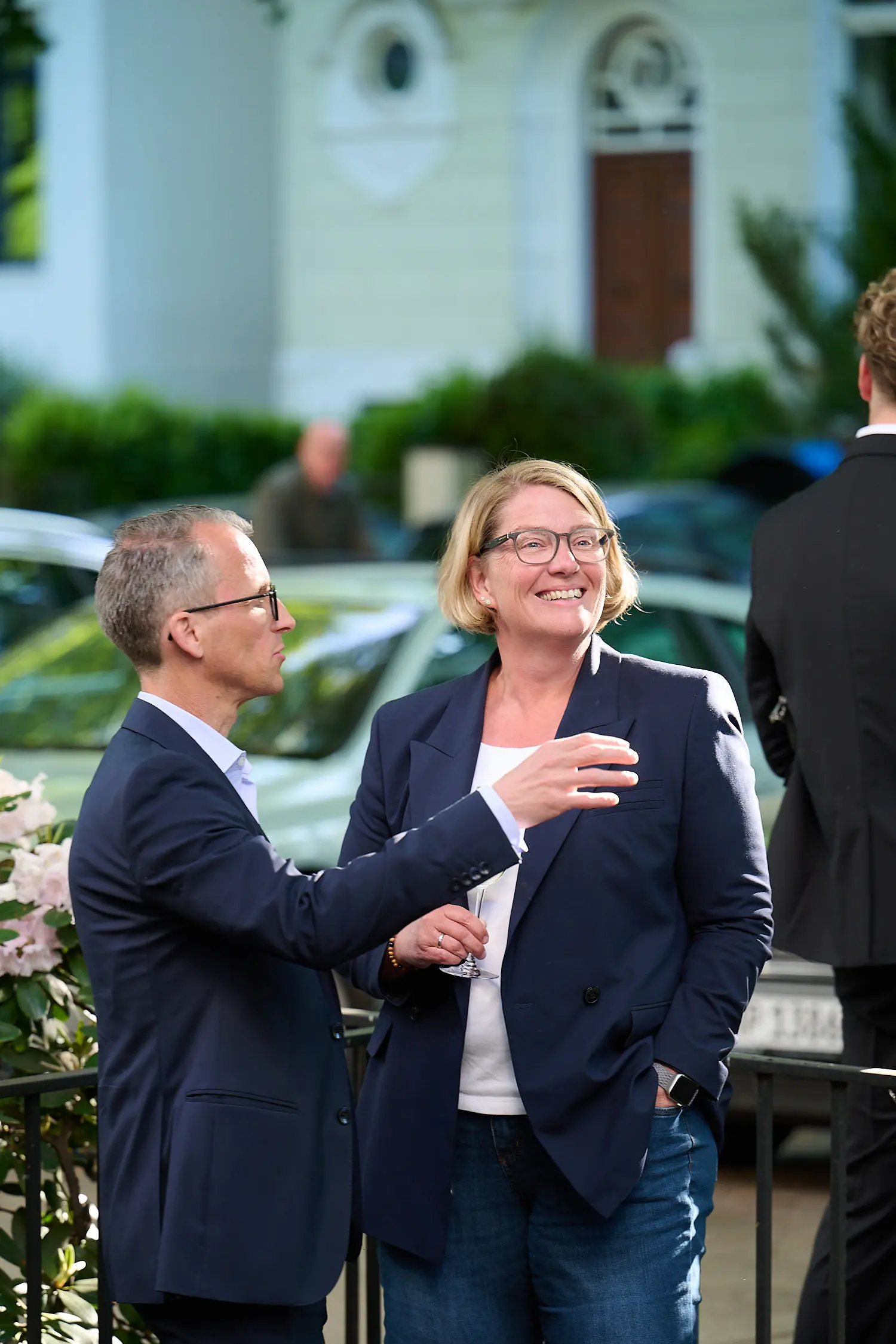 Two professionally dressed people smiling and talking outdoors near a vehicle and flowers.