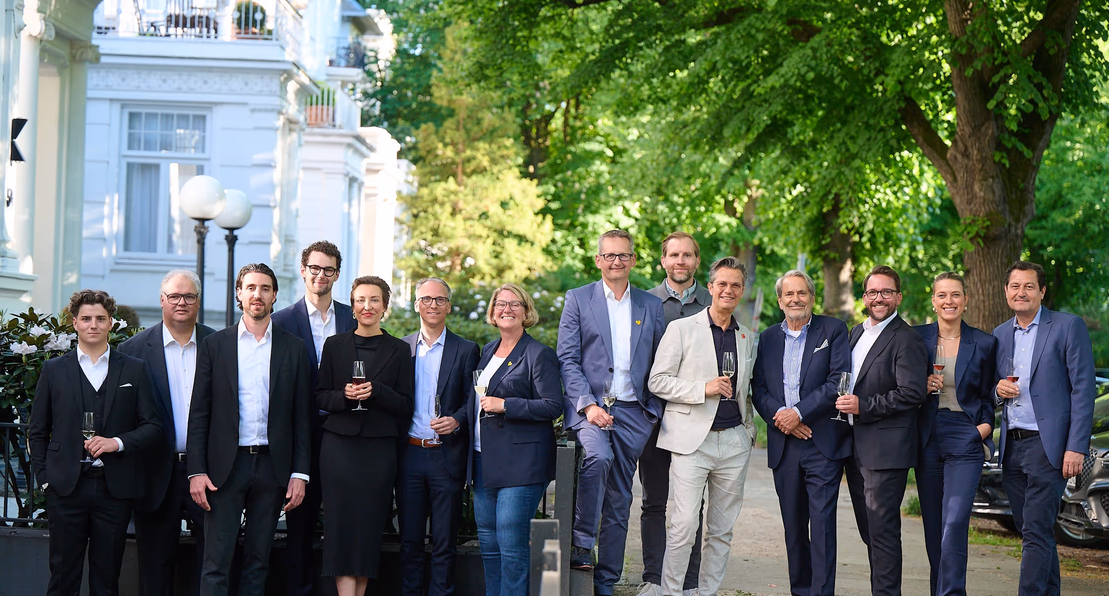 Group of fifteen professionally dressed people standing outdoors with drinks in hand, smiling under green leafy trees.