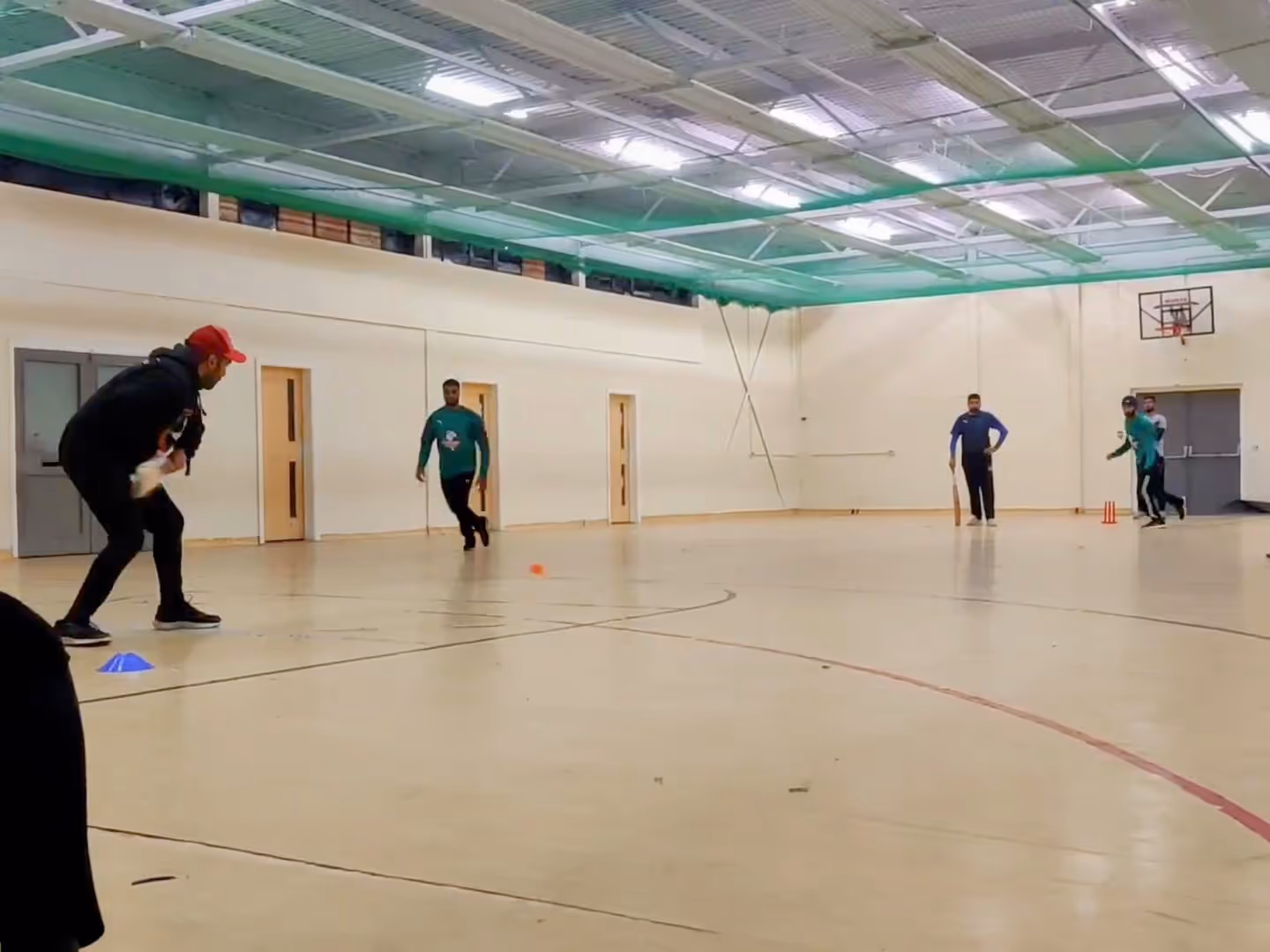 Full indoor cricket action at Yorkshire Muslim Academy (YMA) during the Cricket Arena Men’s Mini Bash League 2021 — batter prepares to play as bowler delivers and fielders move in mid-play.