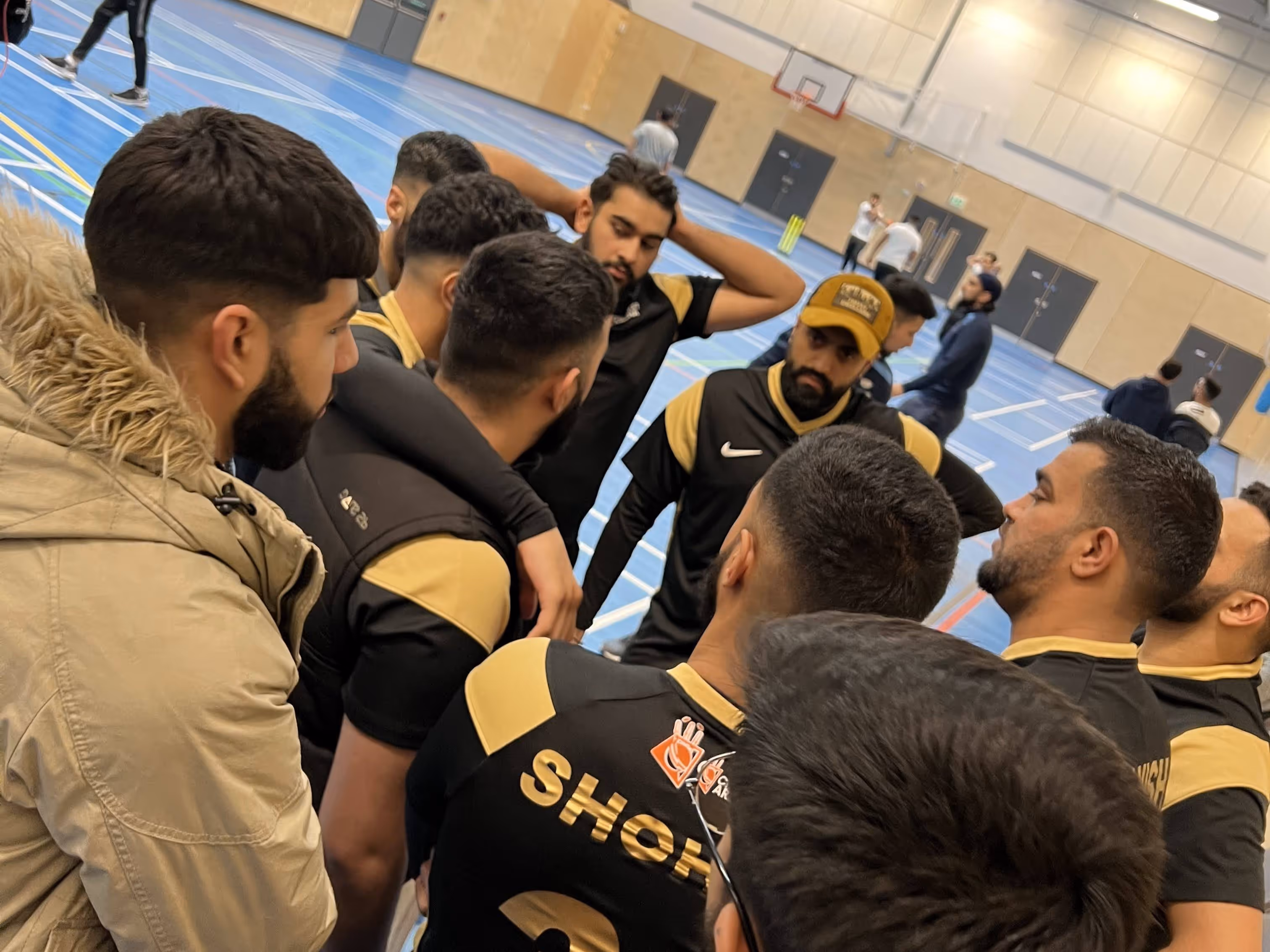 Sheffield Darnall Dynamites team gathered in a focused huddle before the Yorkshire Indoor Finals match against Bradford, discussing tactics and batting order in their black and gold Cricket Arena kits at the indoor venue.