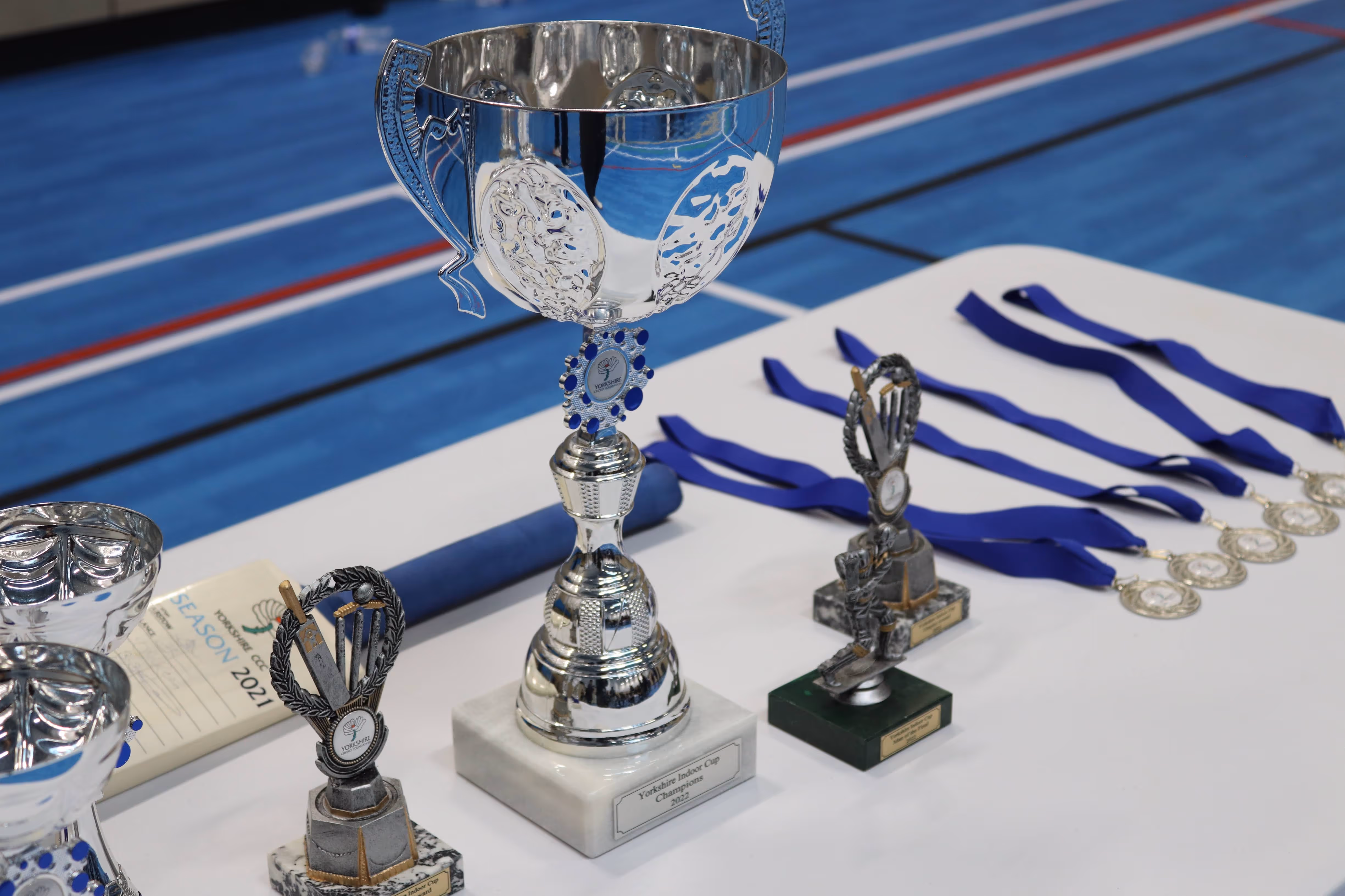 Display of awards and trophies on a presentation table, including the Yorkshire Indoor Cities Cup champions trophy, medals, player awards, and a cricket bat signed by Yorkshire players, ready for the ceremony.