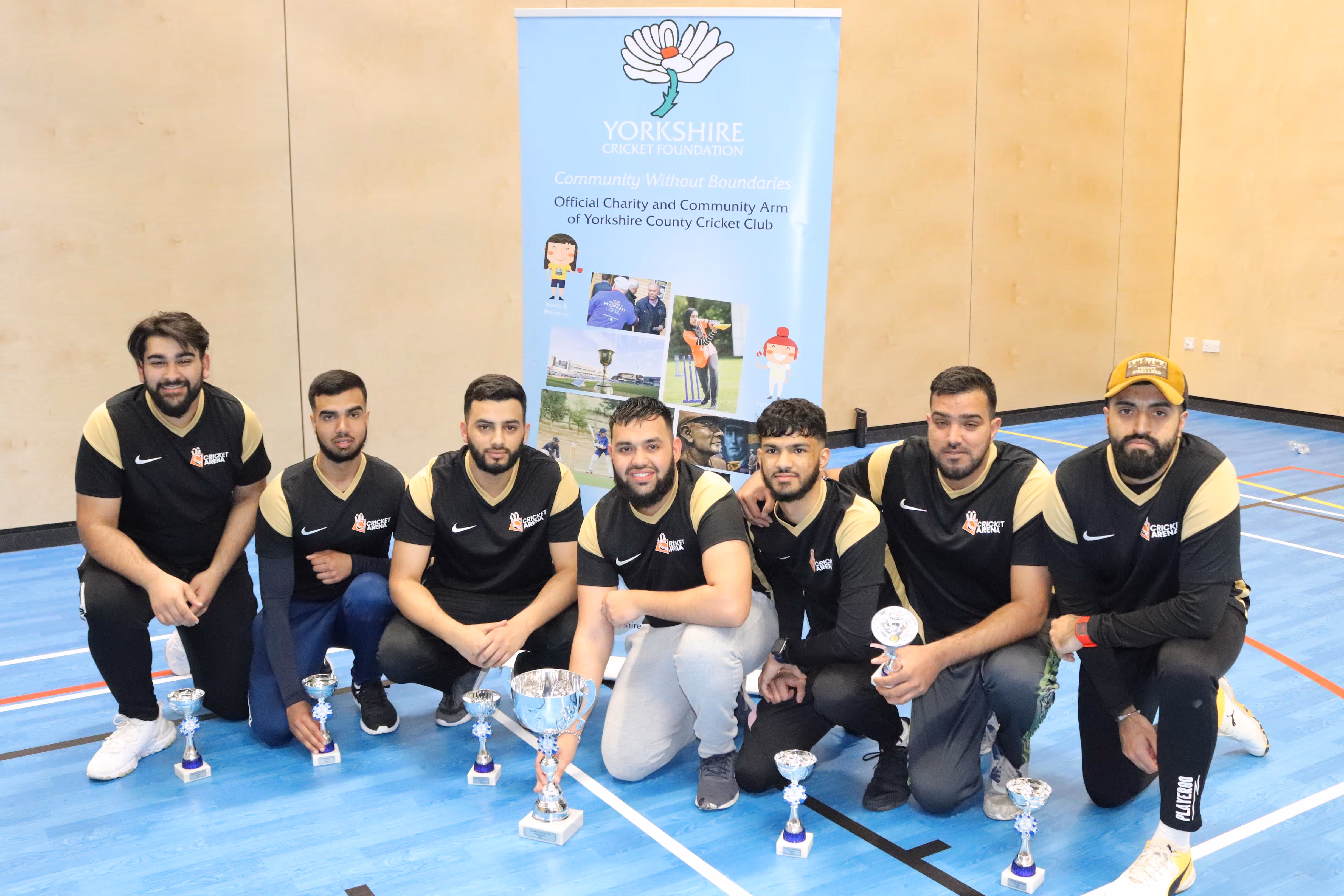 Sheffield Darnall Dynamites team kneeling proudly with their trophies after the Yorkshire Indoor Finals presentation, wearing black and gold Cricket Arena kits with the Yorkshire Cricket Foundation banner displayed behind them.