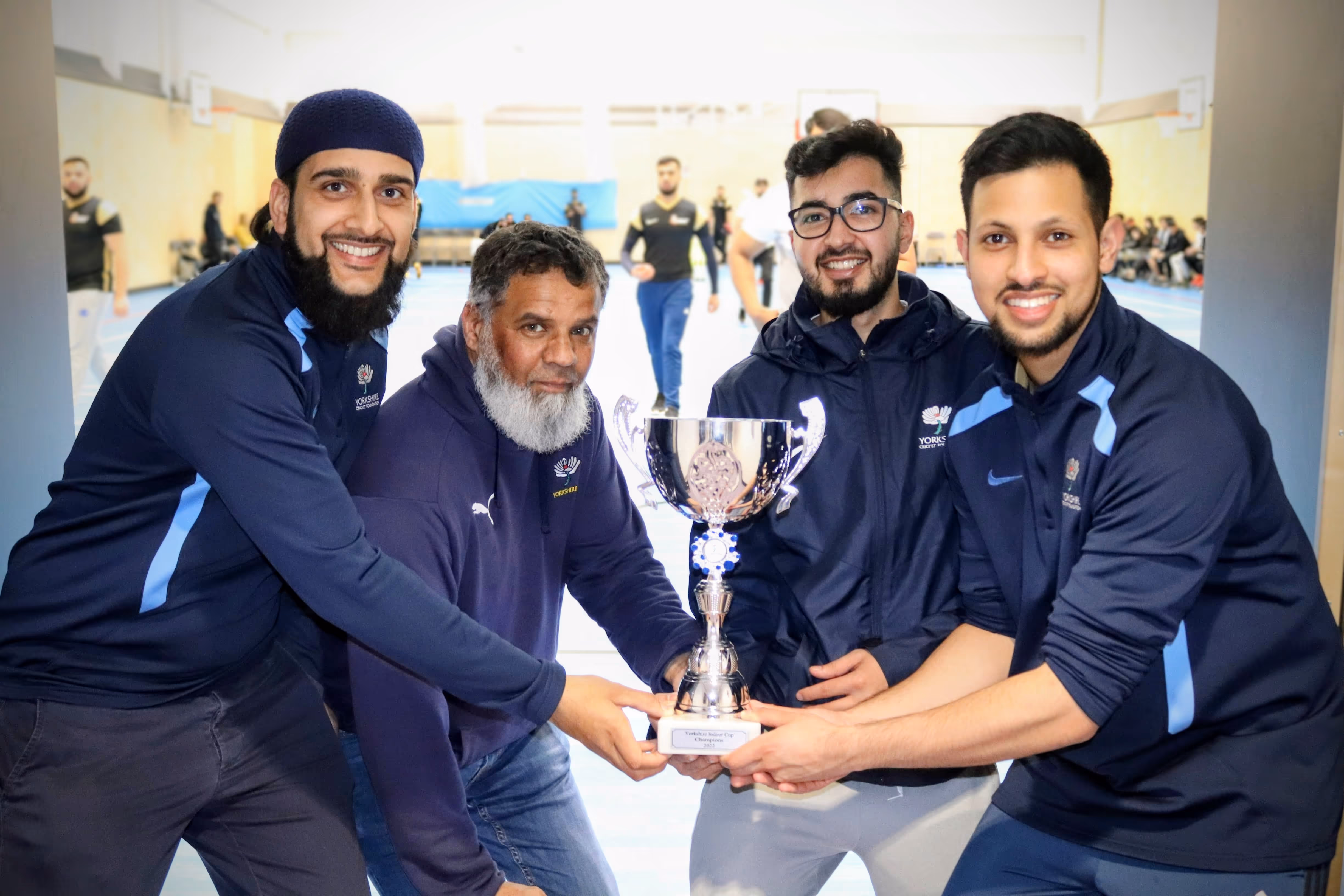 Yorkshire Cricket Foundation staff — Hamzah Hussain, Sohail Raz, Soyeb Hayat and Zain ul Abdin — proudly holding the Yorkshire Indoor Cup trophy together, with indoor cricket action visible in the background at New College Bradford.