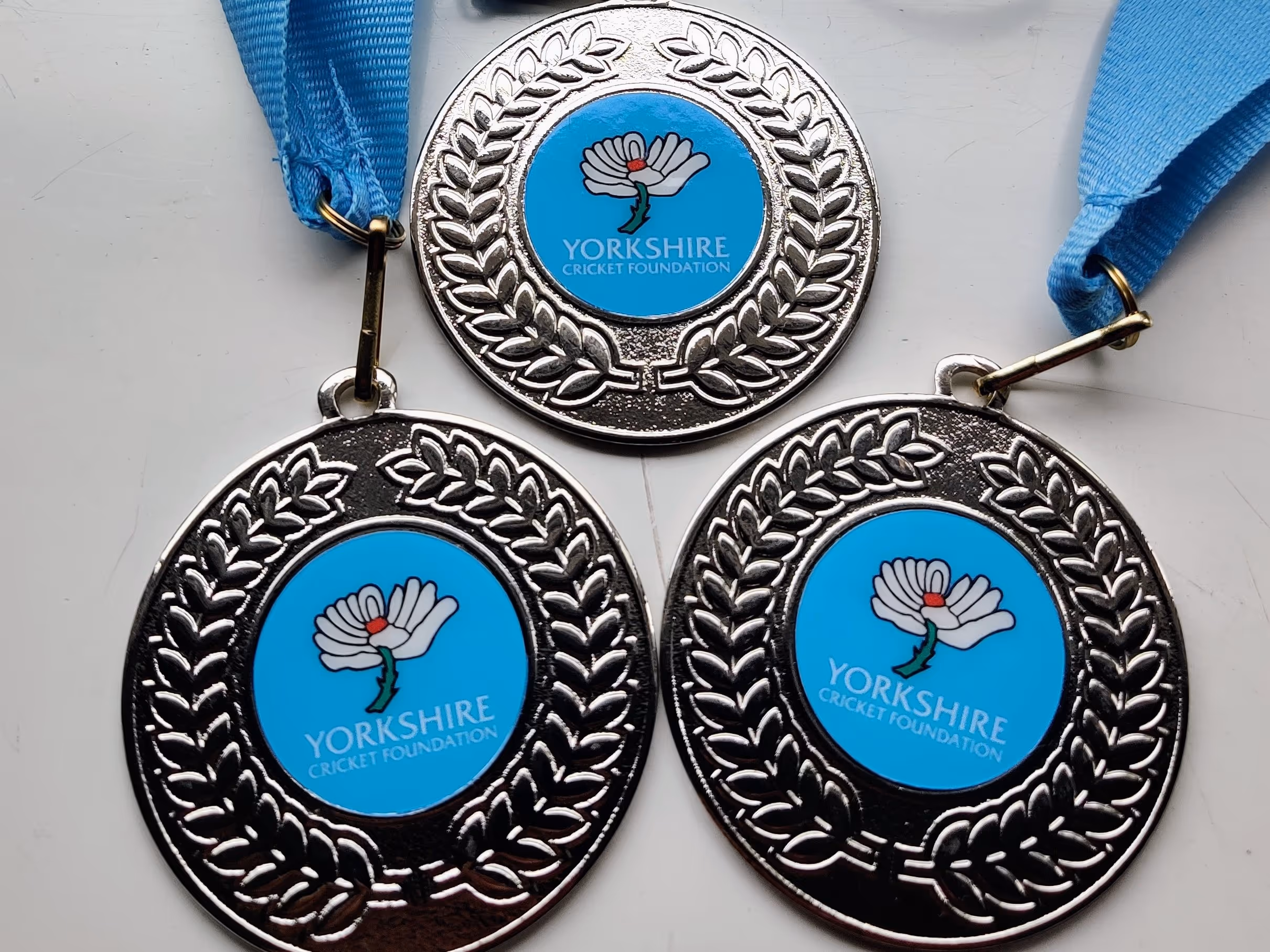 Close-up of three silver participation medals with the Yorkshire Cricket Foundation logo. Each medal symbolises friendship, confidence, and skills gained through taking part in the Junior Hundred 2022 community cricket event.