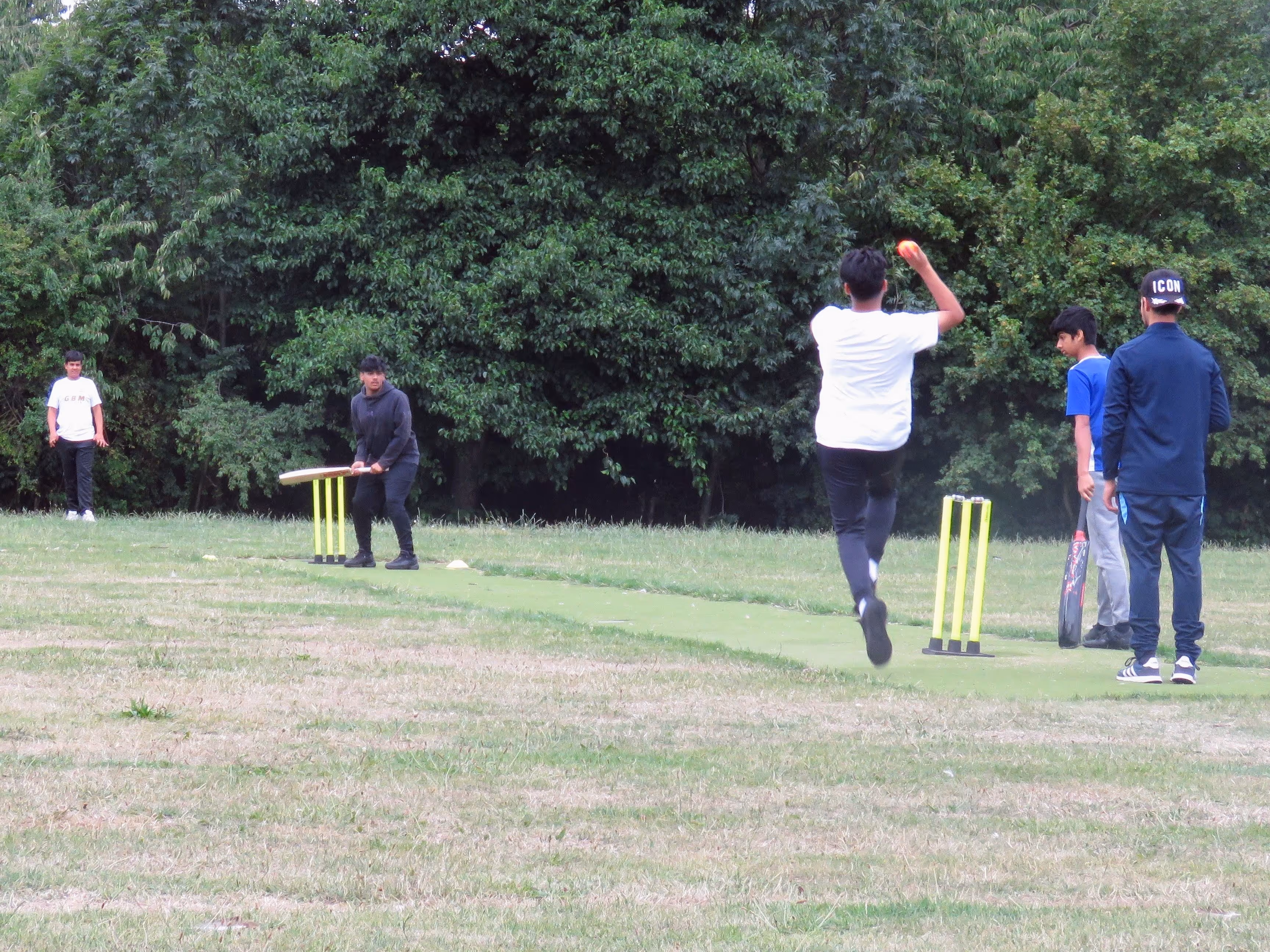 Creative action photo from the Junior Hundred 2022 showing batter Haseeb playing a skilled shot as fielders prepare to stop the ball during a fun windball cricket match on a non-turf pitch at Kashmir Gardens.