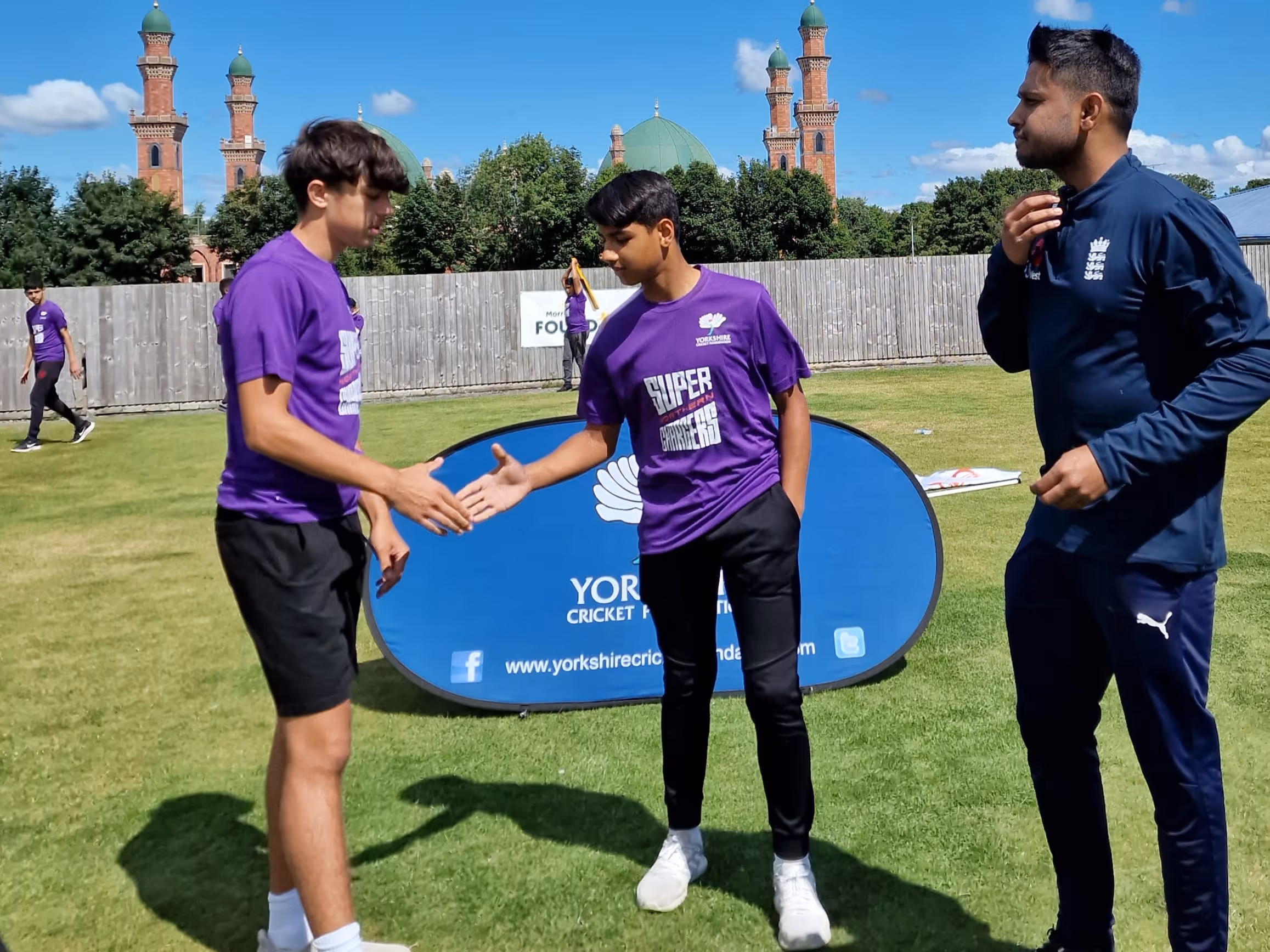 Captains Rizwan and Lyle shake hands at the toss before the Junior Hundred final at Park Avenue, Bradford, with the YCF banner and mosque domes in the backdrop.