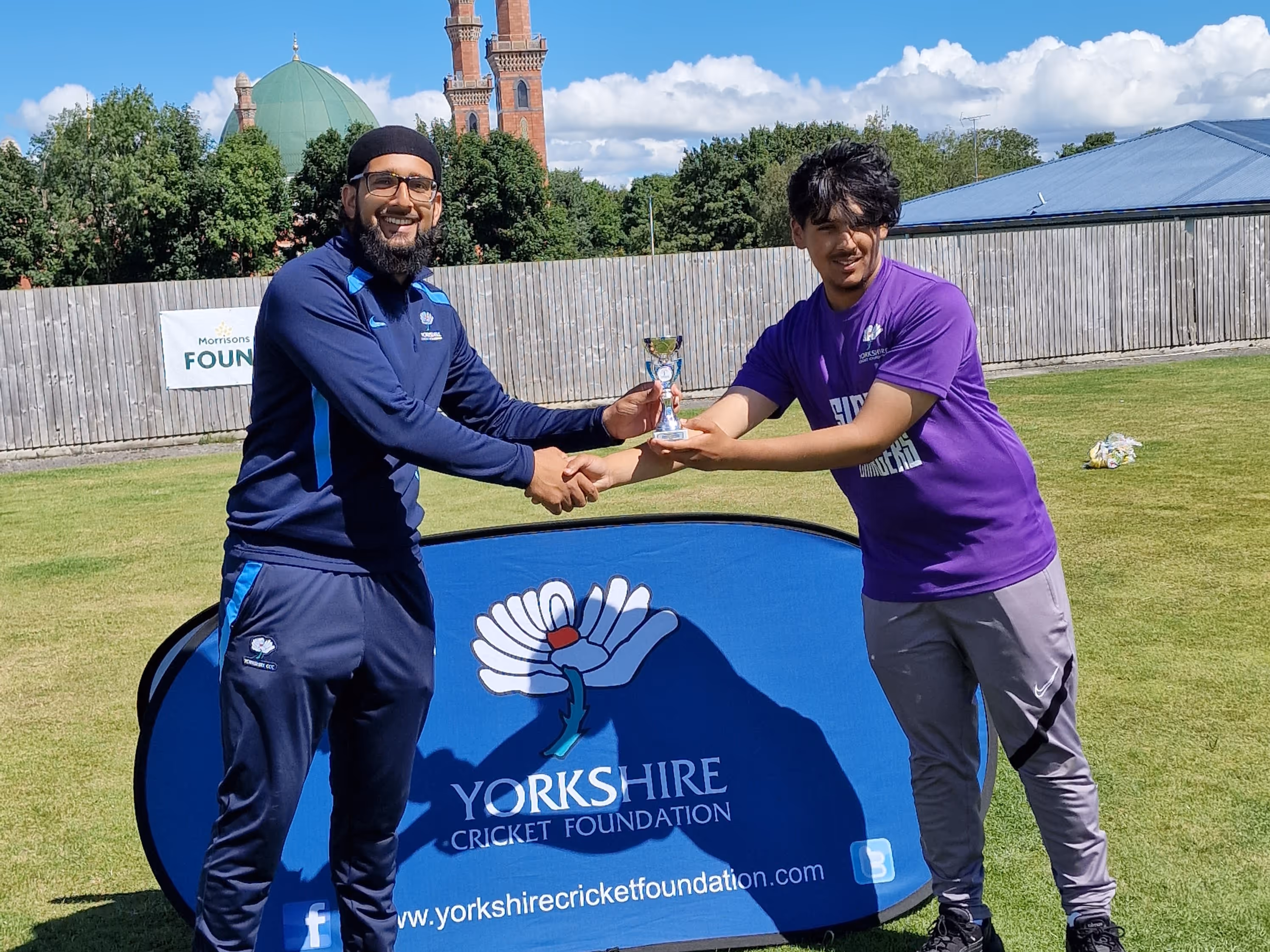 Top batter Hasan receives his award from Sohail Raz in front of the Yorkshire Cricket Foundation banner, with the mosque domes visible in the Bradford backdrop.
