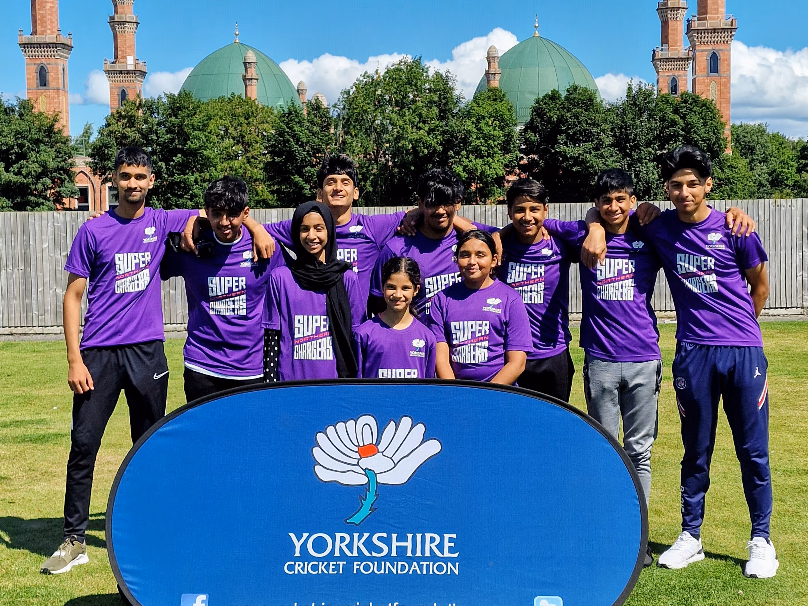 Sheffield’s Burngreave Tigers team pose before their Junior Hundred 2022 match at Park Avenue, Bradford. Boys and girls stand arm in arm behind the YCF banner with the mosque backdrop.