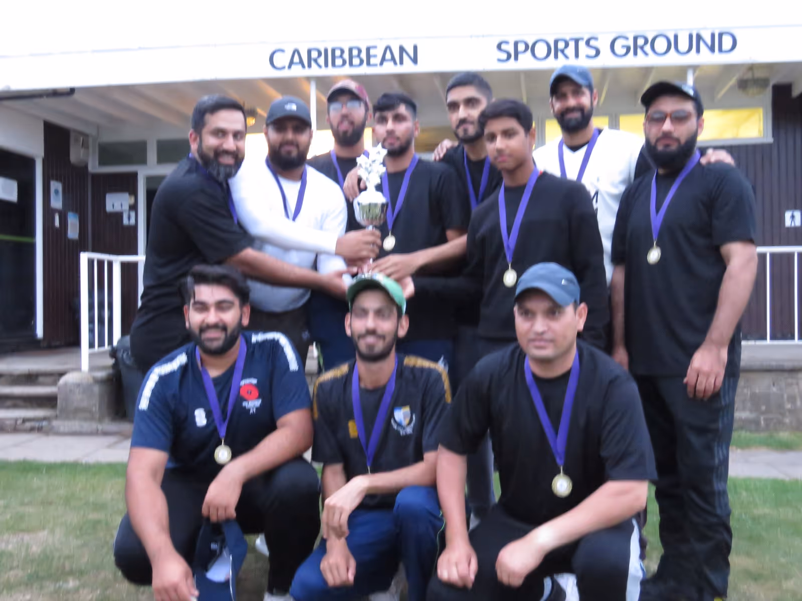 The Firth Park Warriors team pose together with the winning trophy outside the Caribbean Sports Club pavilion — smiles, medals, and unity after their league triumph.