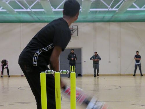 Indoor cricket action at YMA sports hall as bowler runs in and batter prepares, showcasing South Asian community sport