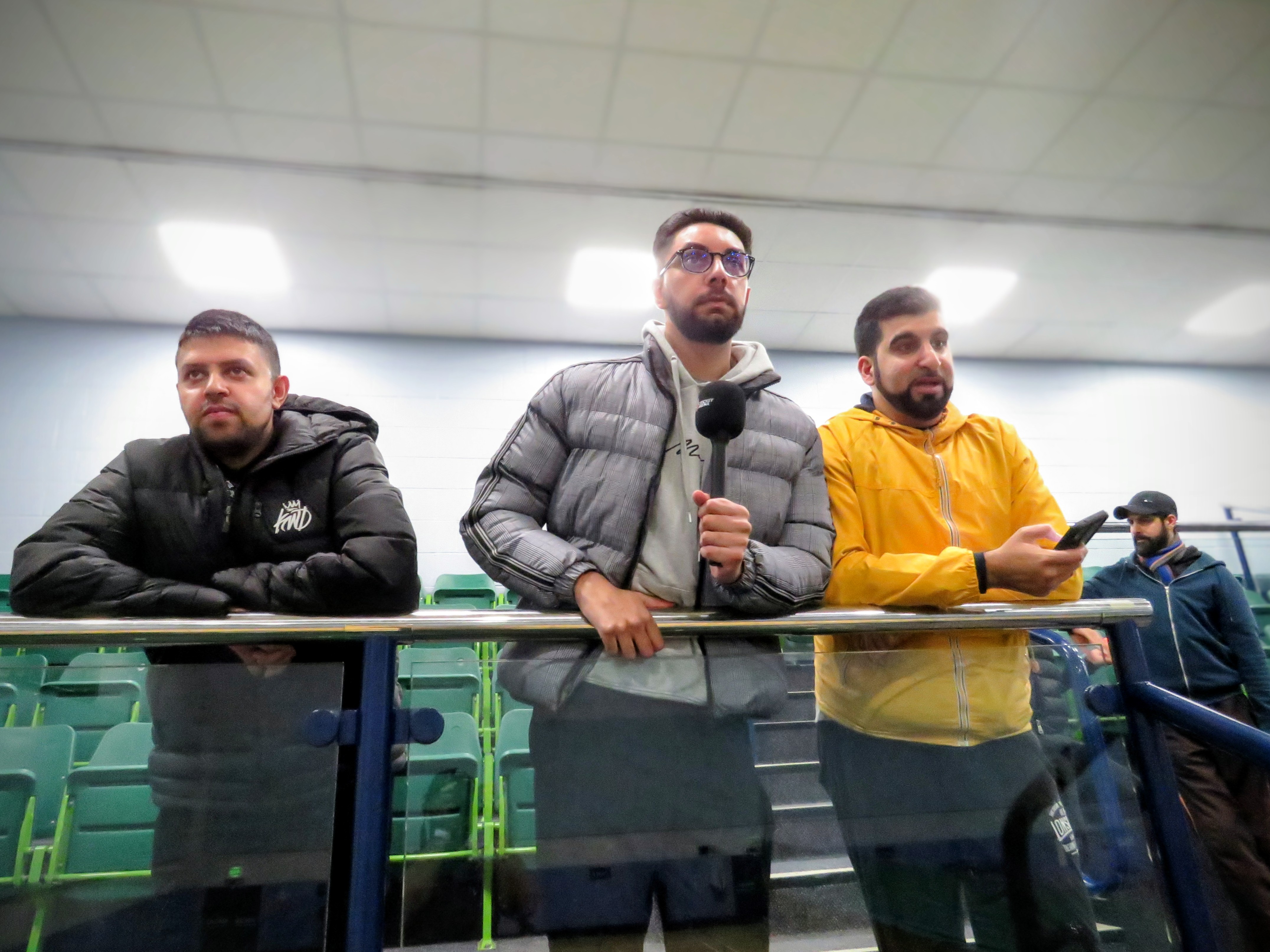 Volunteer commentators on the balcony at Hillsborough Leisure Centre holding a Cricket Arena microphone and checking live CricClubs scores during an indoor match.
