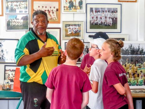 Milton Samuels passionately sharing Windrush stories with school children inside the Caribbean Sports Club, surrounded by historic cricket photos celebrating Caribbean heritage.