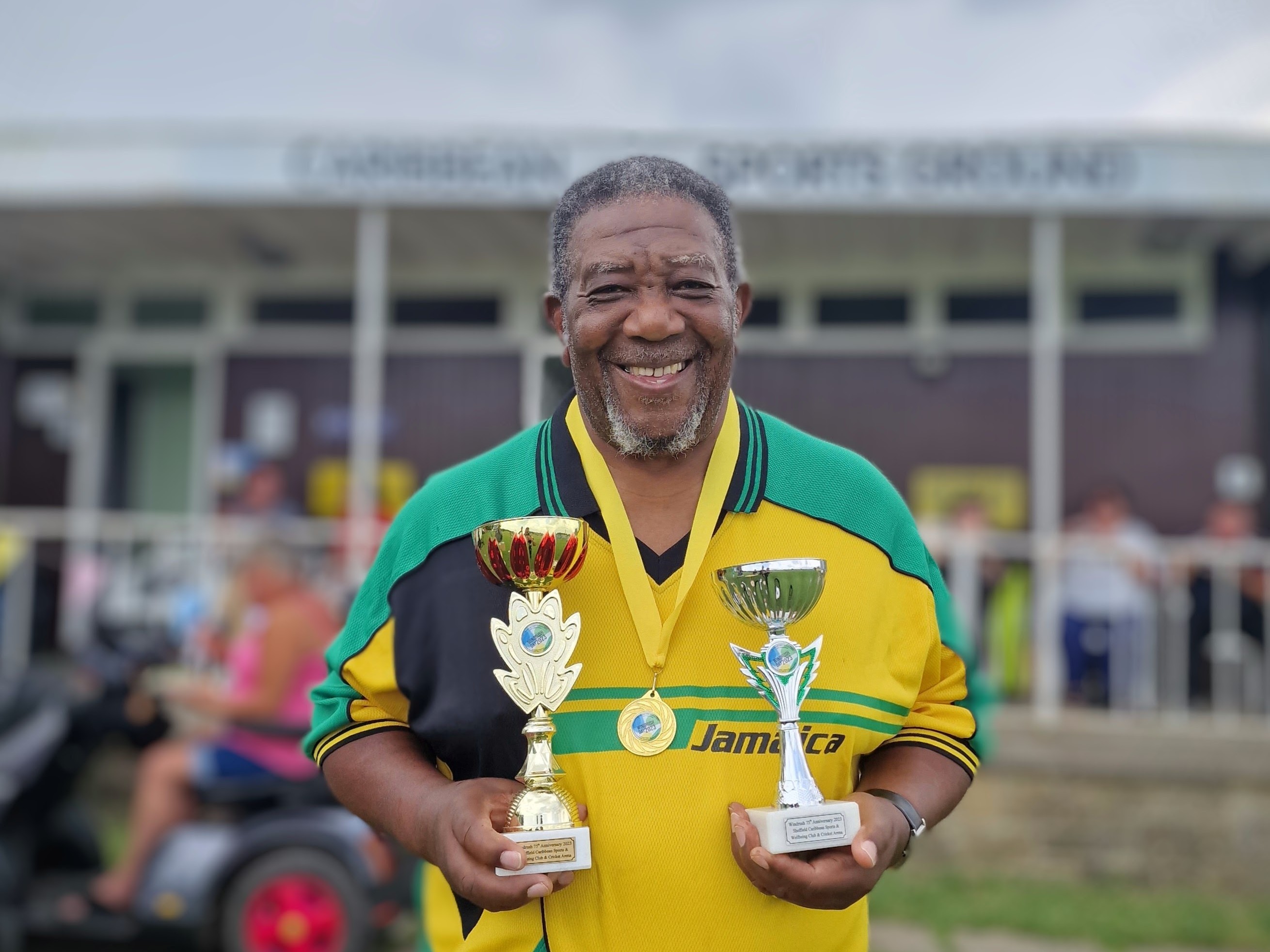 Coach Milton Samuels proudly holds two winning trophies and wears a medal, smiling in front of the Sheffield Caribbean Sports Club pavilion after the Windrush75 celebration event.
