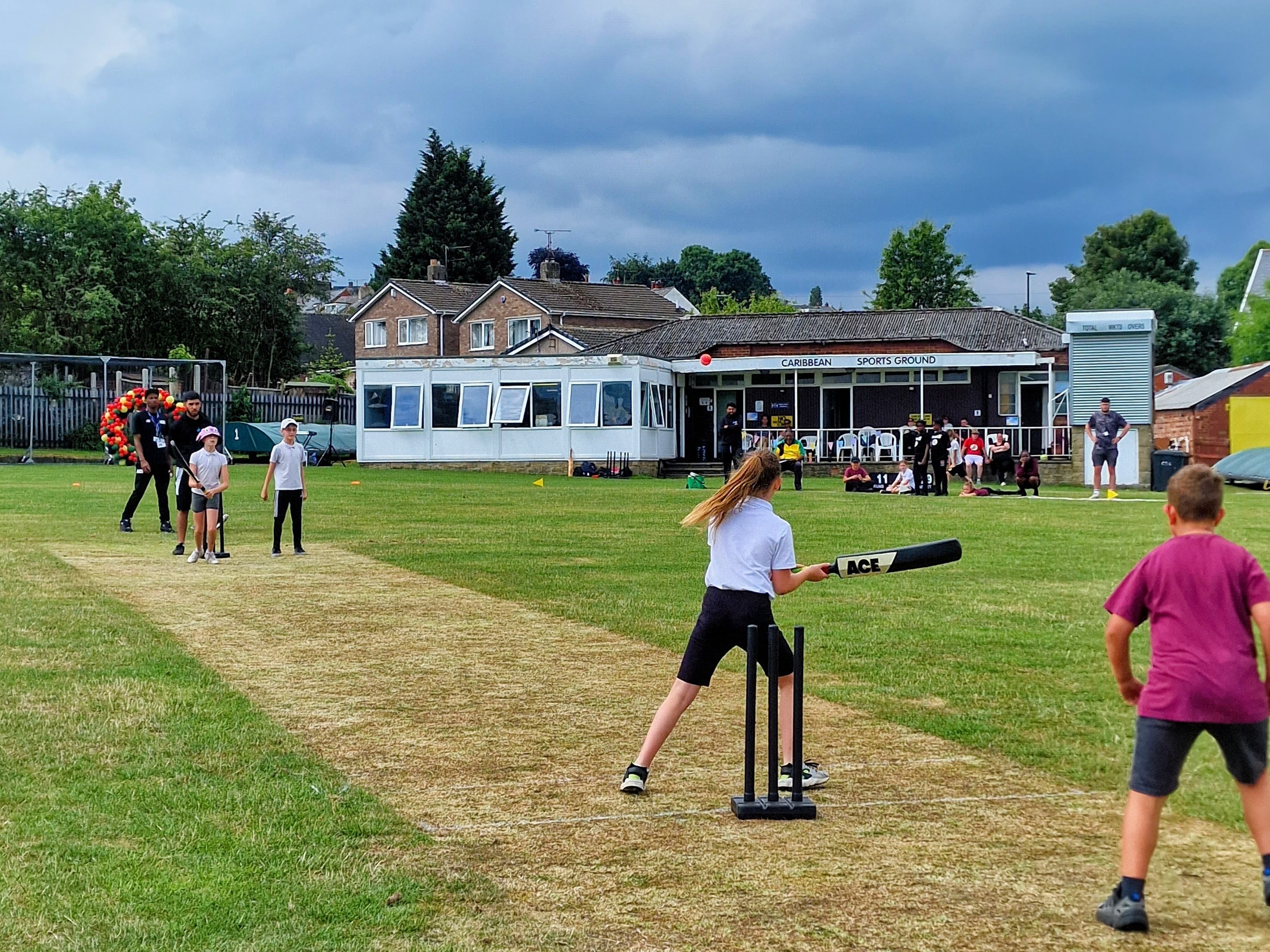 Young girl plays a cricket shot using an ACE cricket bat on the grass wicket at Sheffield Caribbean Sports Club, with the pavilion and community coaches in the background.