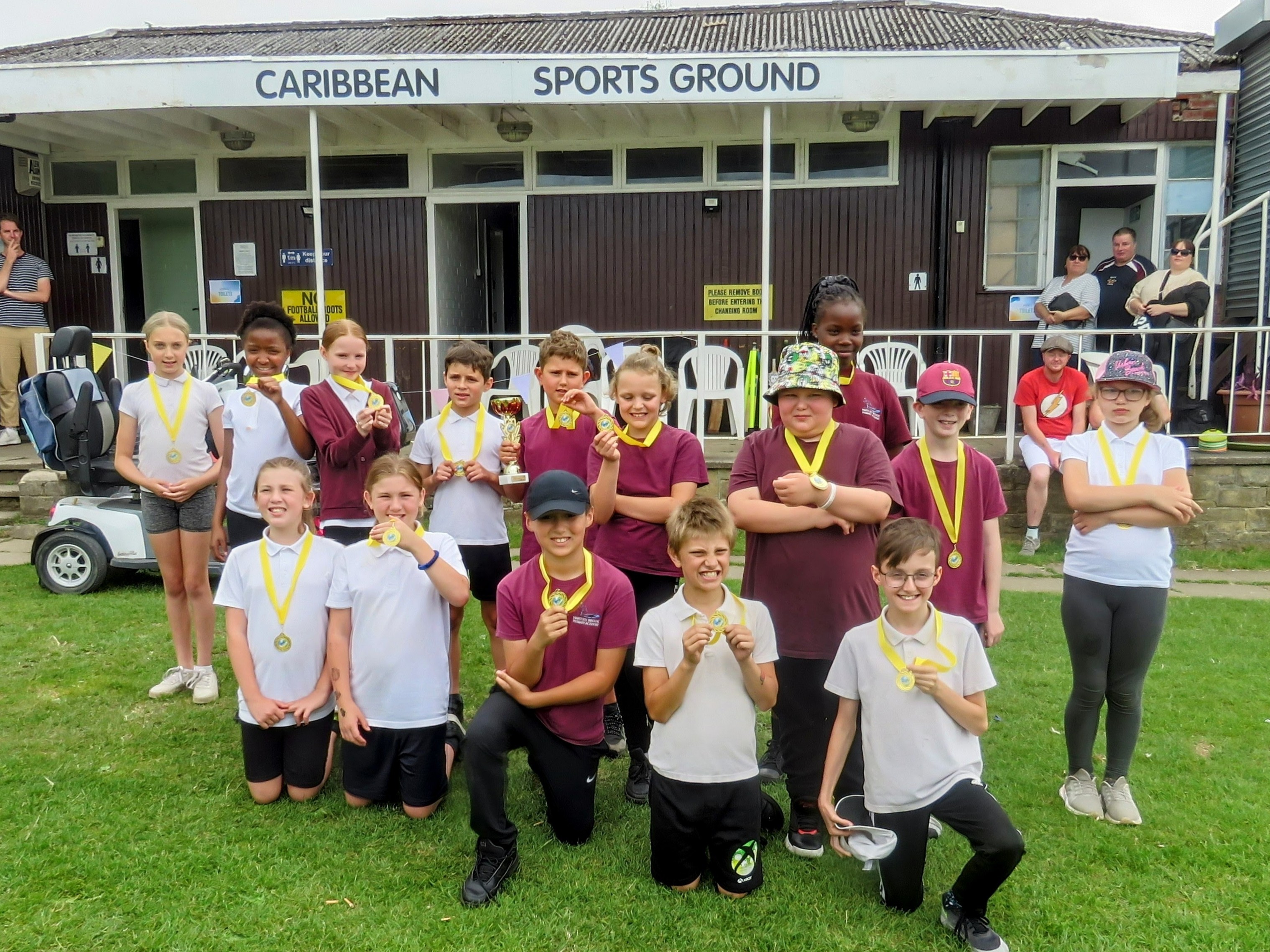 roup photo of all the school children posing proudly with their medals and trophy in front of the Sheffield Caribbean Sports Club pavilion after the Windrush75 celebration.
