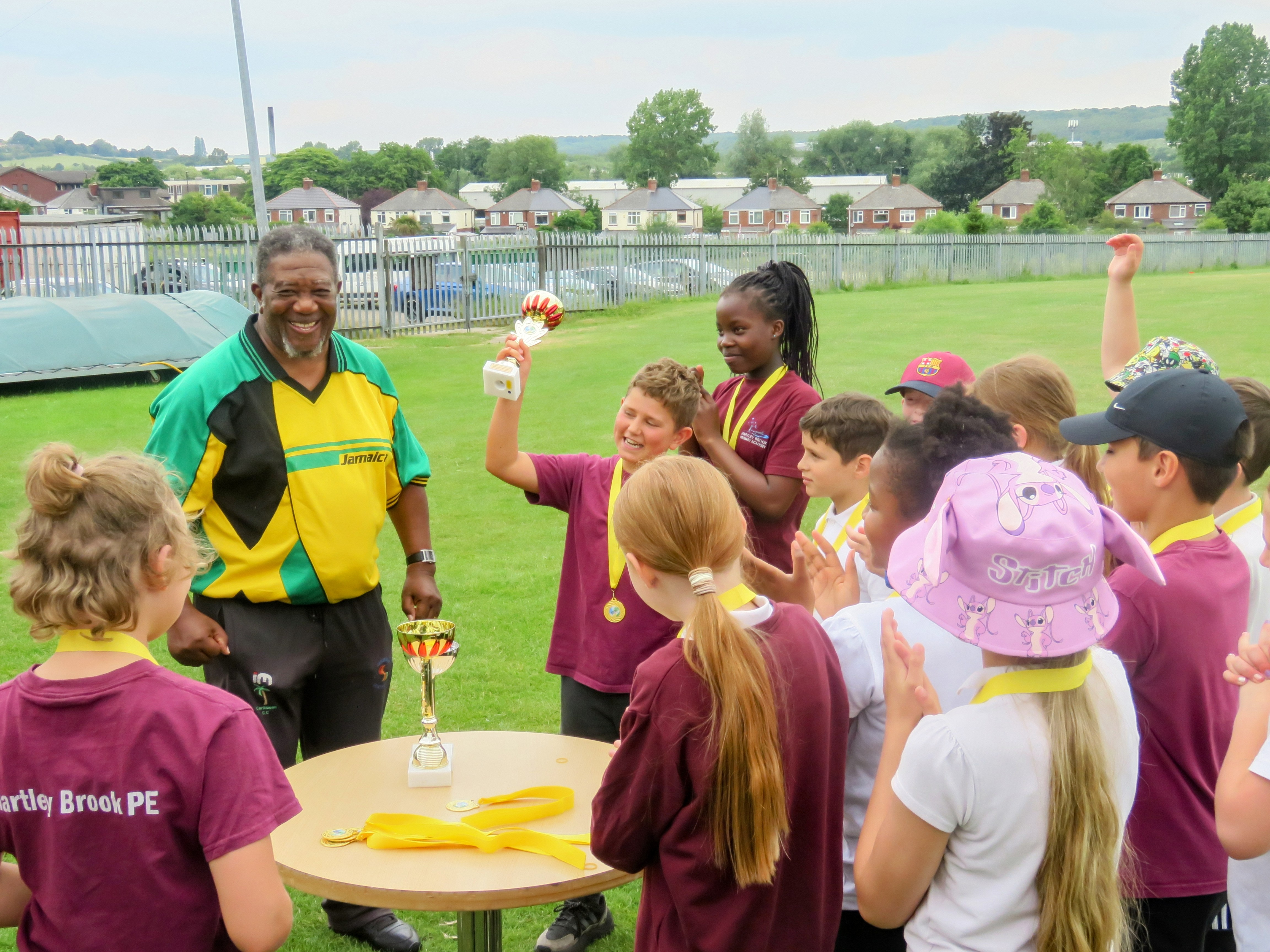 A smiling Milton Samuels stands proudly as a young pupil lifts the winning trophy during the Windrush75 presentation at Sheffield Caribbean Sports Club, surrounded by classmates wearing medals.