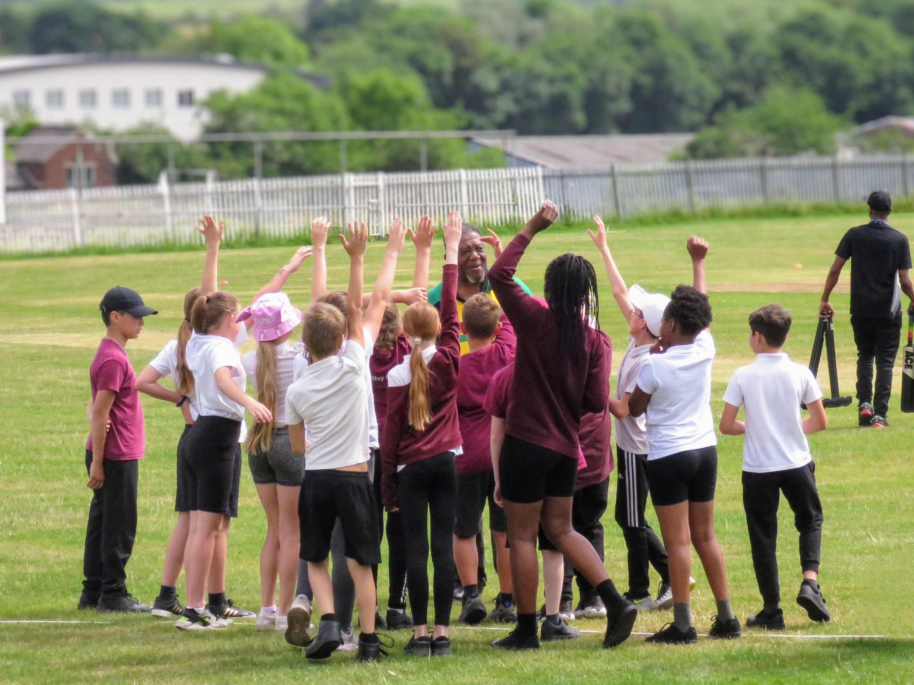 A lively group of school children cheer with their hands raised as coach Milton Samuels engages them during a fun Windrush75 cricket session at Sheffield Caribbean Sports Club.