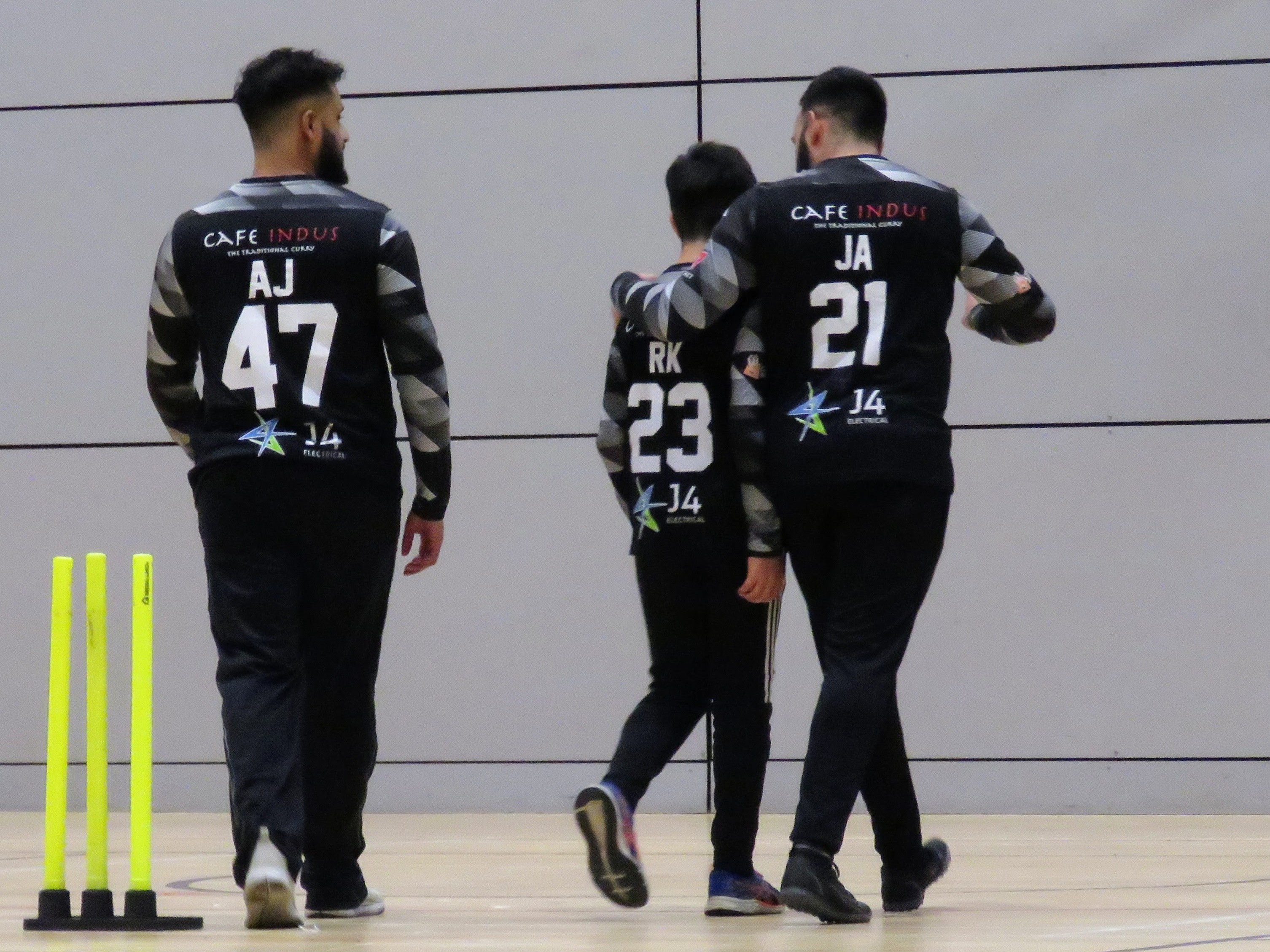 Young bowler Rehan Khan walks back supported by senior teammate Jawad Akhtar during the BBICL final, highlighting youth development and mentoring. The backs of their kits display team sponsors Café Indus and J4 Electrical.