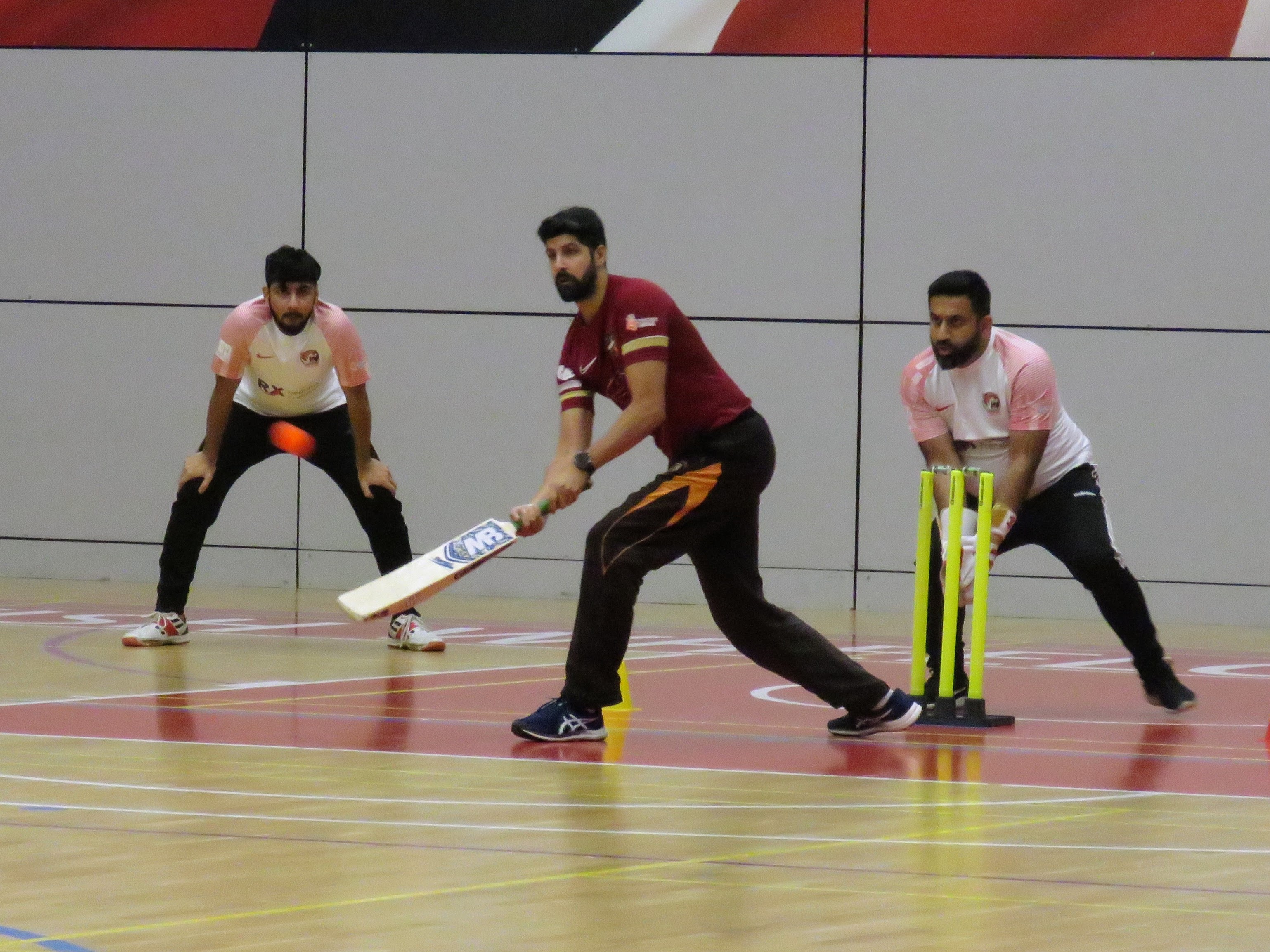 Haidar Rasool plays an innovative scoop shot during an indoor windball cricket match at EIS basketball hall, with Awais positioned at slip and Adeel wicketkeeping close behind.