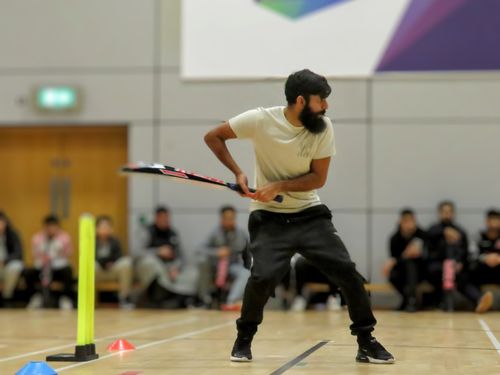Irfan prepares to play a powerful shot during an indoor windball match at EIS, captured from the leg-umpire’s view with a fibreglass bat in hand and a crowd of spectators watching from the benches in the background.