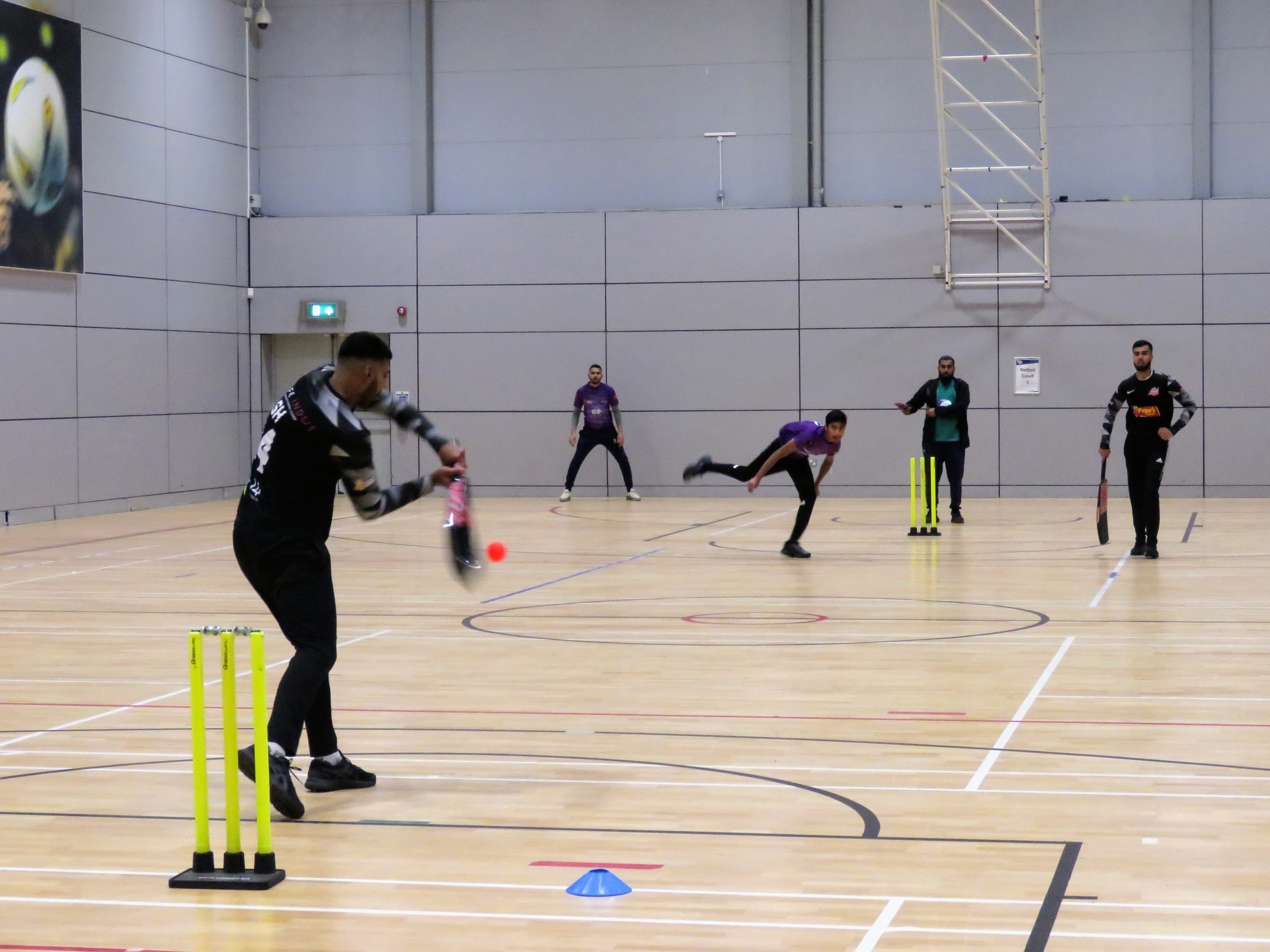 Saqib strikes a big six against Yahya during the BBICL final at the English Institute of Sport, with the ball captured mid-flight just before connecting with the bat. The scene shows all players in view, highlighting the intensity of the moment