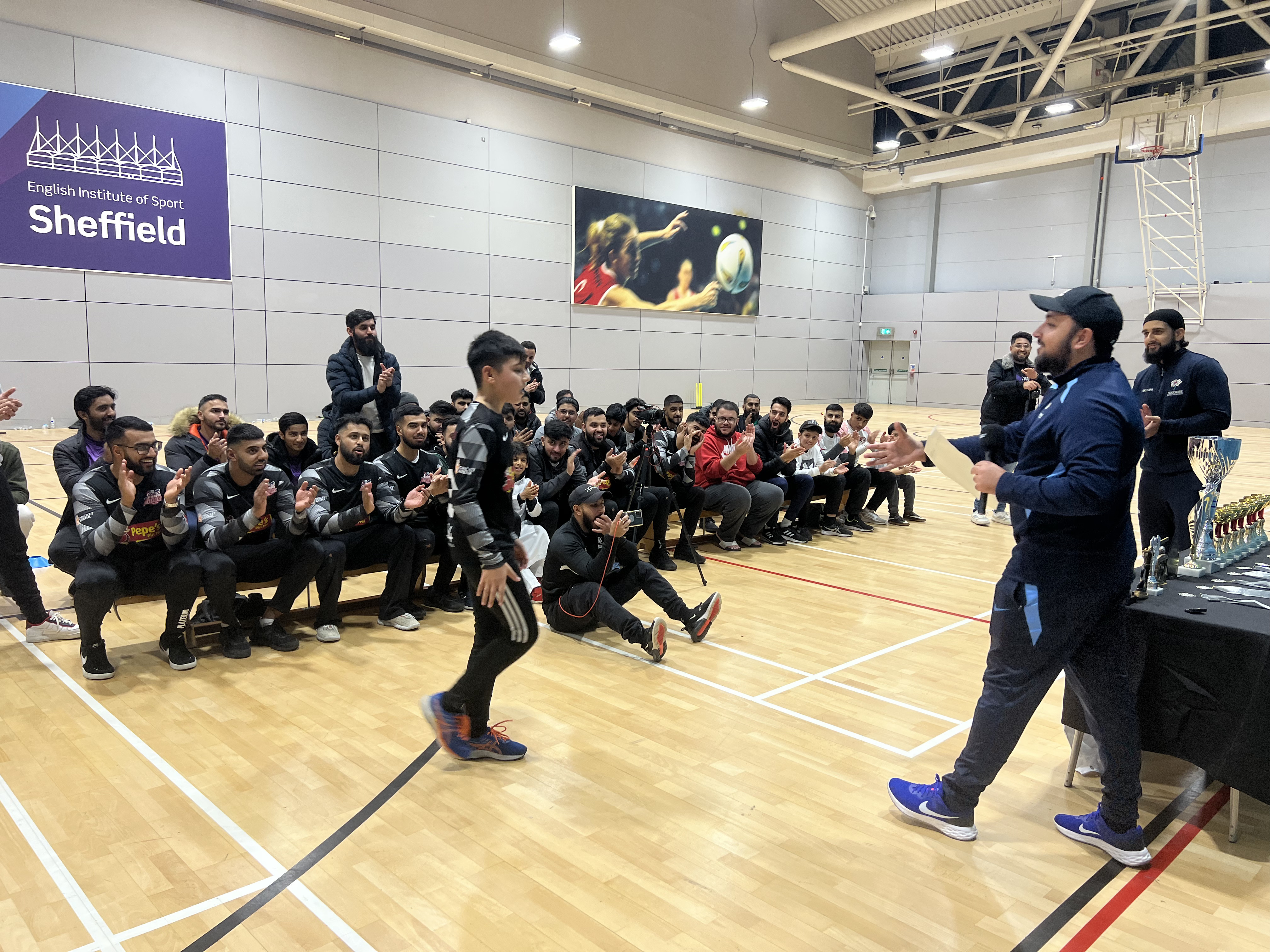 Spectators and participants fill three rows of benches, clapping as young player Rehan Khan walks up to receive his Man of the Final award at EIS. A proud moment that captures Cricket Arena’s mission — youth development and community support