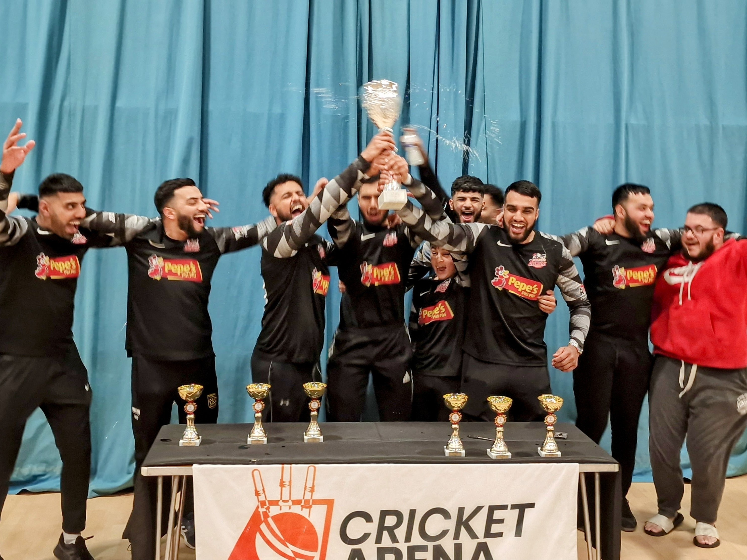 “Pitsmoor Avengers players erupt in joy as they lift the BBICL Champions Trophy at the English Institute of Sport. The team, wearing their black Pepe’s Piri Piri kits, celebrate with unity and excitement in front of the Cricket Arena banner