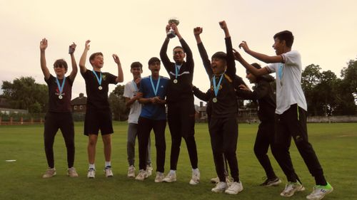The winning team, Sharrow Stars, lift the Junior Hundred 2023 trophy high in celebration — teammates jump and cheer on the lush green field at Shiregreen Cricket Club, all wearing medals and smiling with pride.