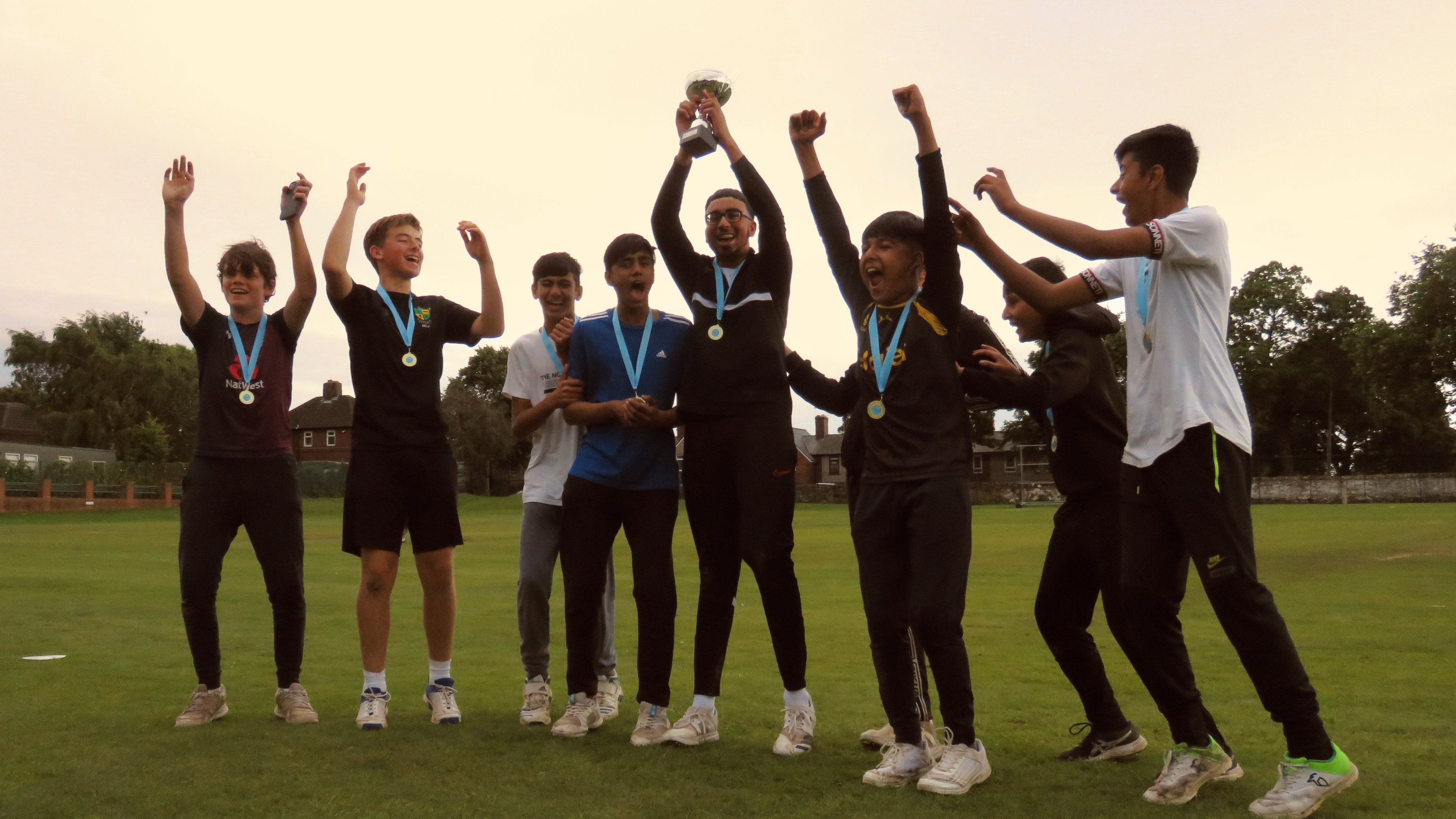 The winning team, Sharrow Stars, lift the Junior Hundred 2023 trophy high in celebration — teammates jump and cheer on the lush green field at Shiregreen Cricket Club, all wearing medals and smiling with pride.