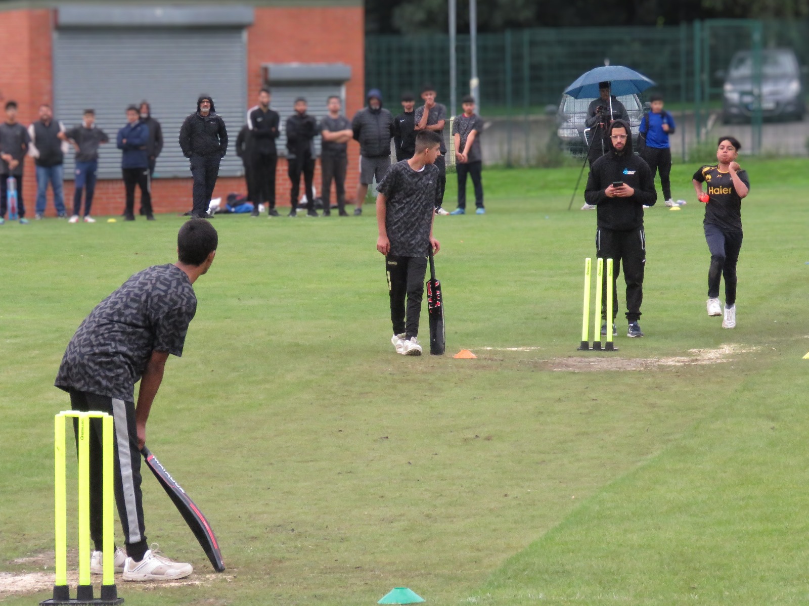 Bowler runs in to deliver as the batter gets ready on the grass wicket — full match action captured with umpire, players, and parents watching on at Shiregreen Cricket Club.