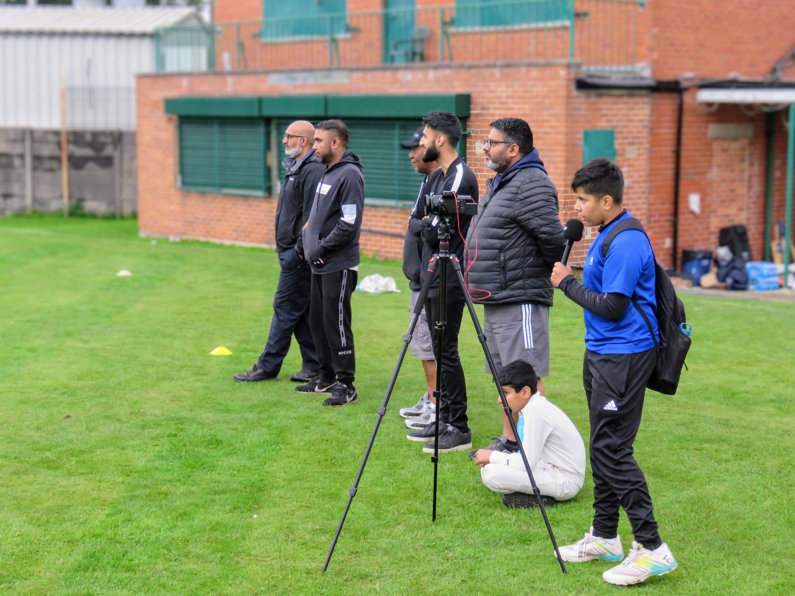 Parents and coaches watch the match while a young volunteer commentates into a microphone beside a camera setup — capturing the game and learning new skills.