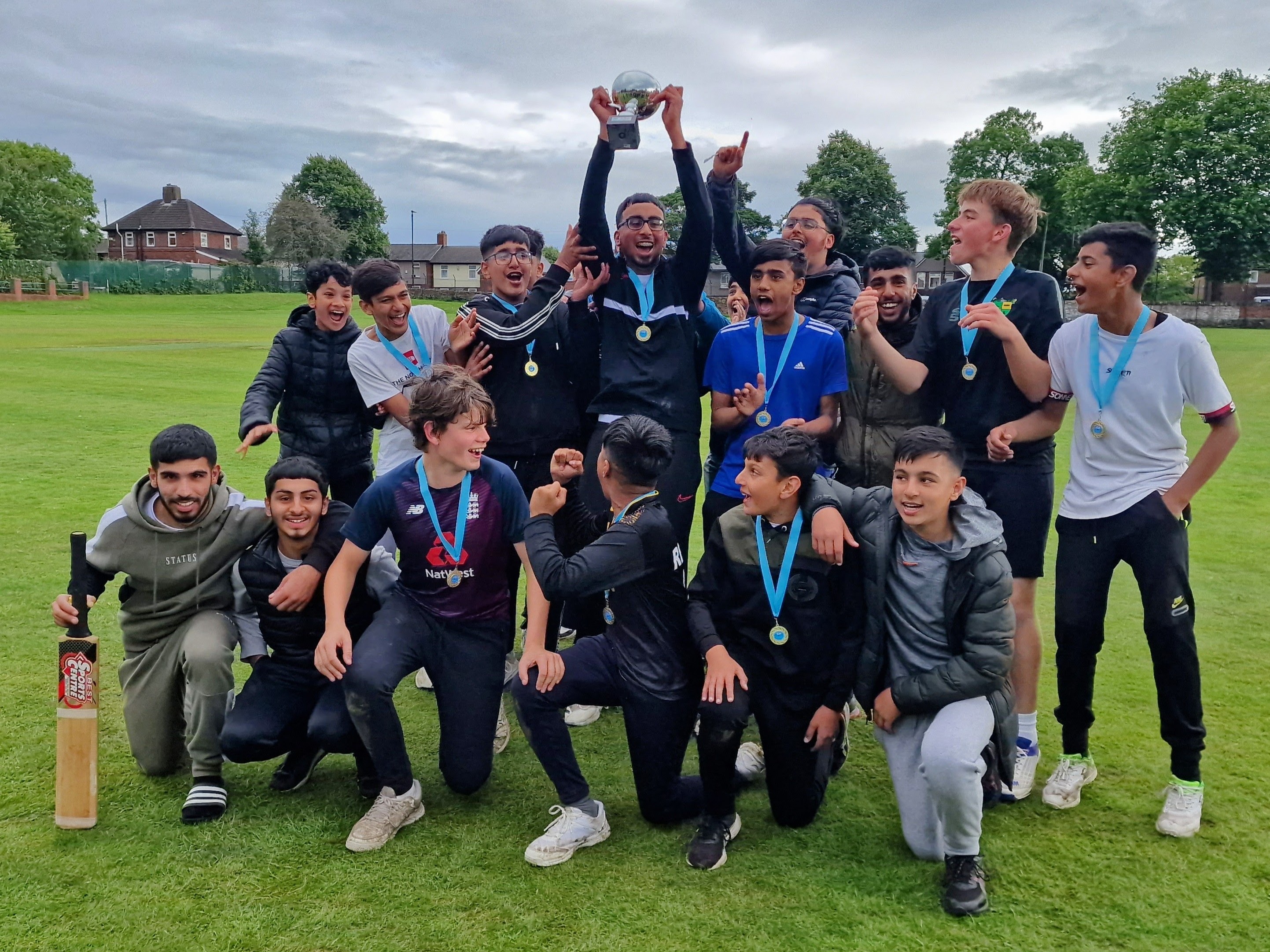 The captain lifts the trophy high surrounded by jubilant teammates and friends, all wearing medals and celebrating victory at the Junior Hundred 2023 presentation at Shiregreen Cricket Club.