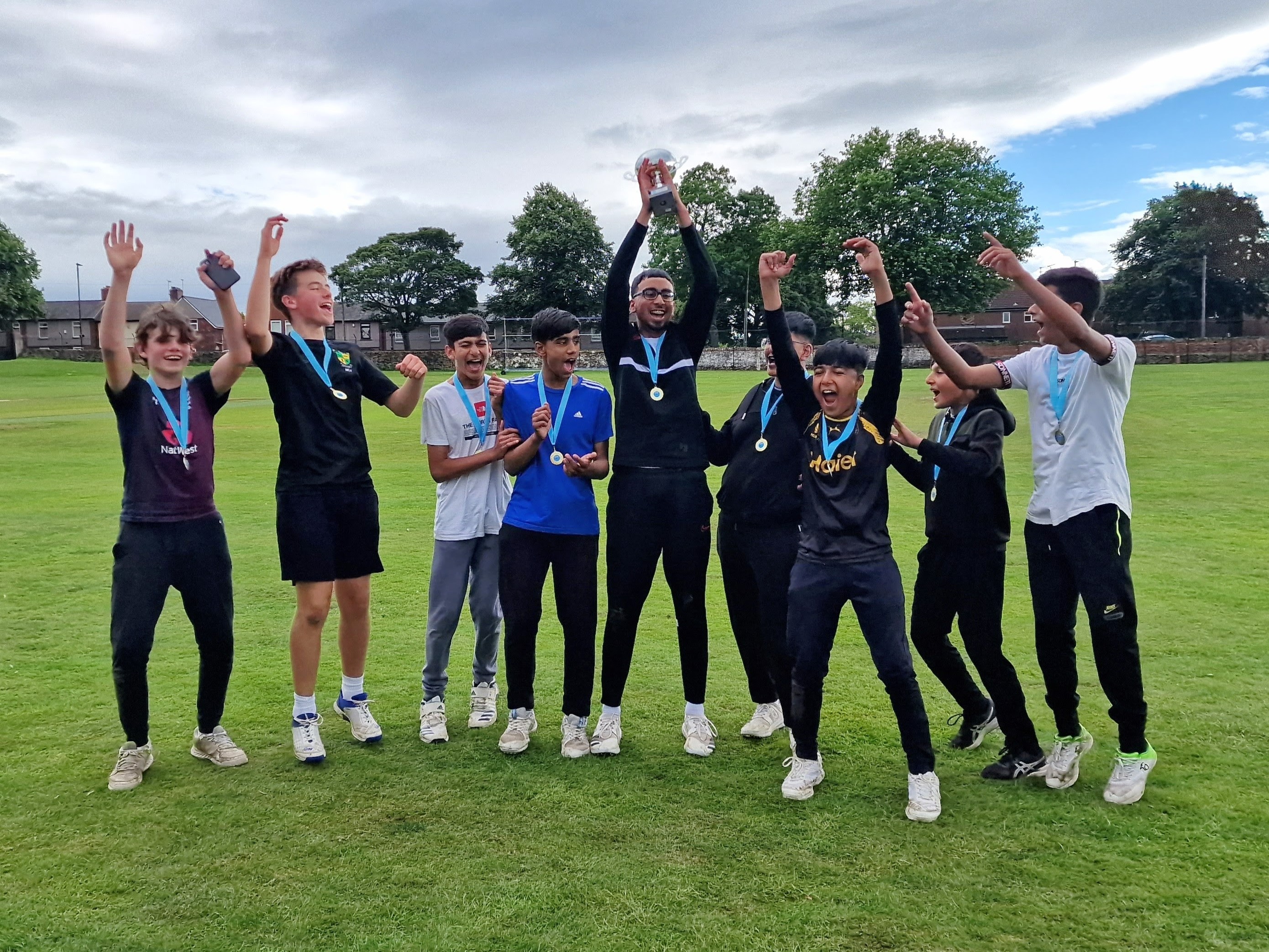 The winning team, Sharrow Stars, lift the Junior Hundred 2023 trophy high in celebration — teammates jump and cheer on the lush green field at Shiregreen Cricket Club, all wearing medals and smiling with pride.