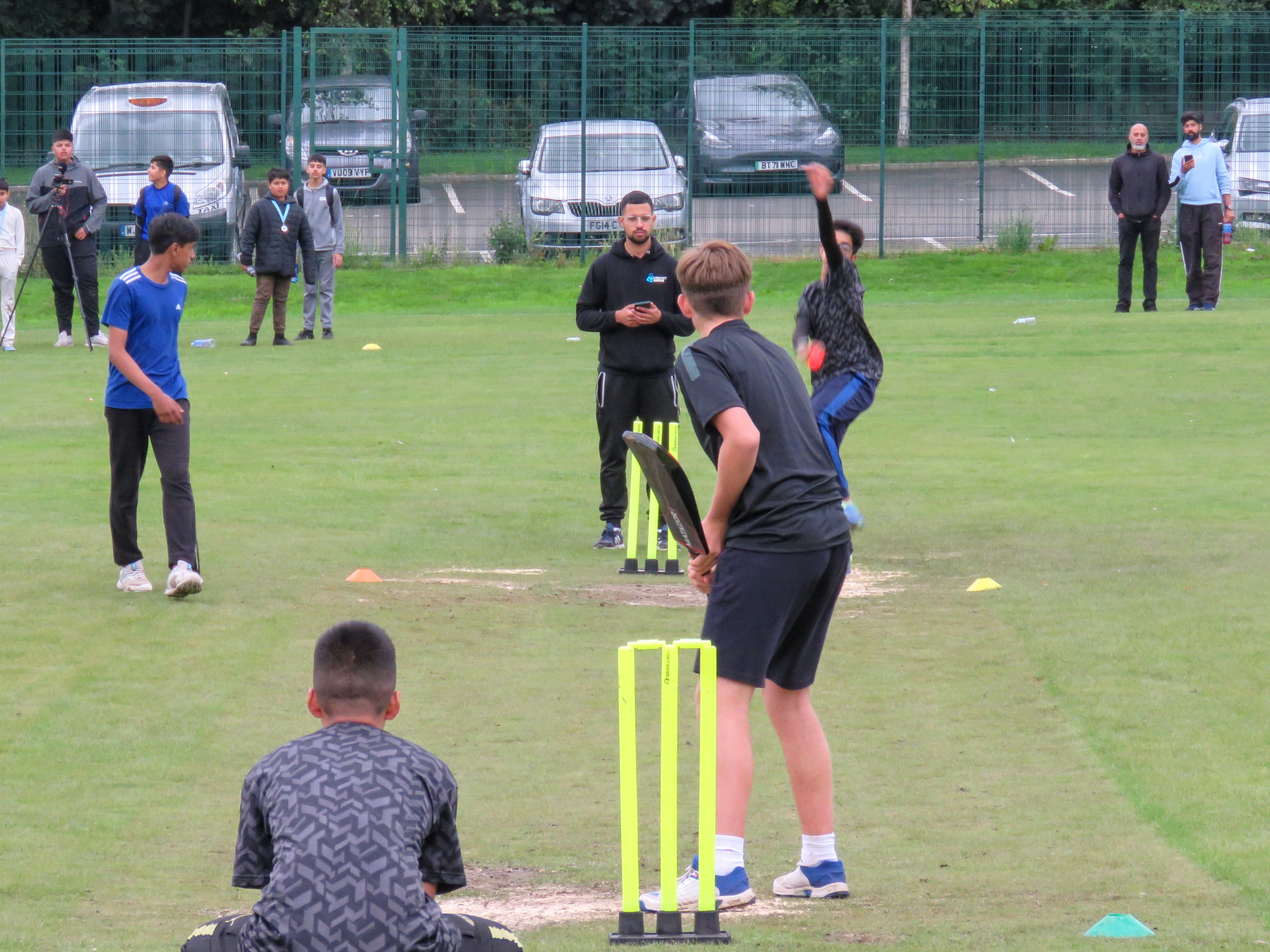 Bowler about to release the ball as a left-handed batter prepares to play his shot on a grass wicket showing sand patches after rain — full match focus with umpire, wicketkeeper, and spectators in view at Shiregreen Cricket Club.