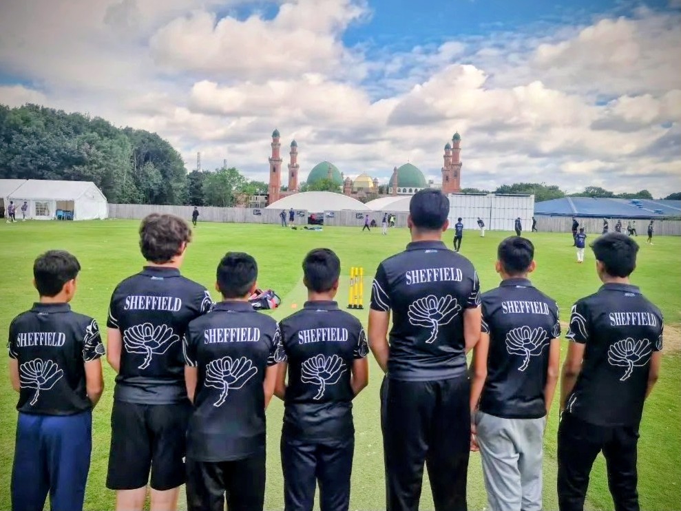 Sheffield team stand on the field with their backs to the camera, showing ‘Sheffield’ and the Yorkshire rose on their shirts as they face the mosque domes at Park Avenue, Bradford during the Junior Hundred 2023.