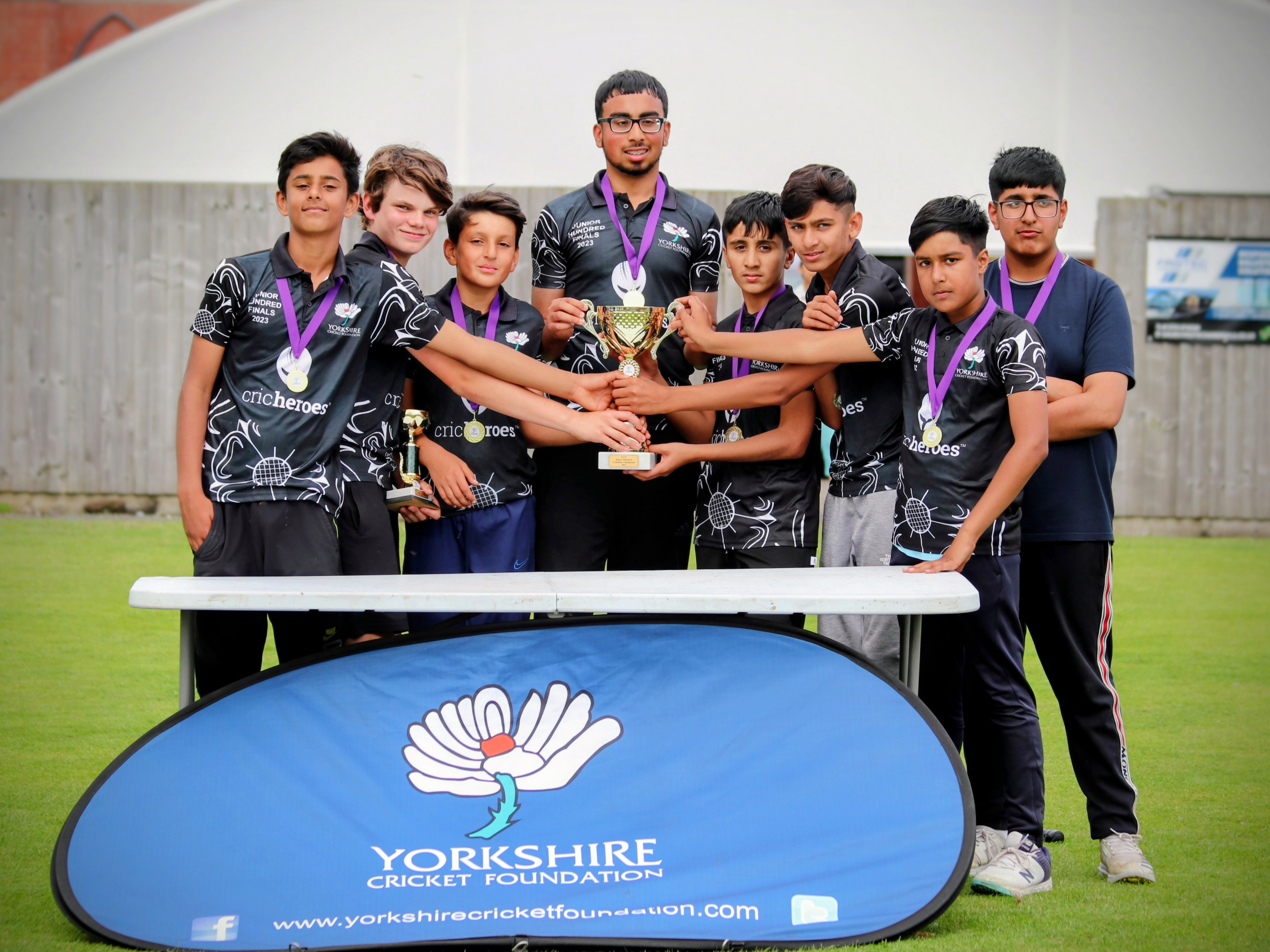 The Cricket Arena Sheffield team celebrate their victory at the Yorkshire Junior Hundred 2023, smiling and holding the trophy together in front of the Yorkshire Cricket Foundation banner.