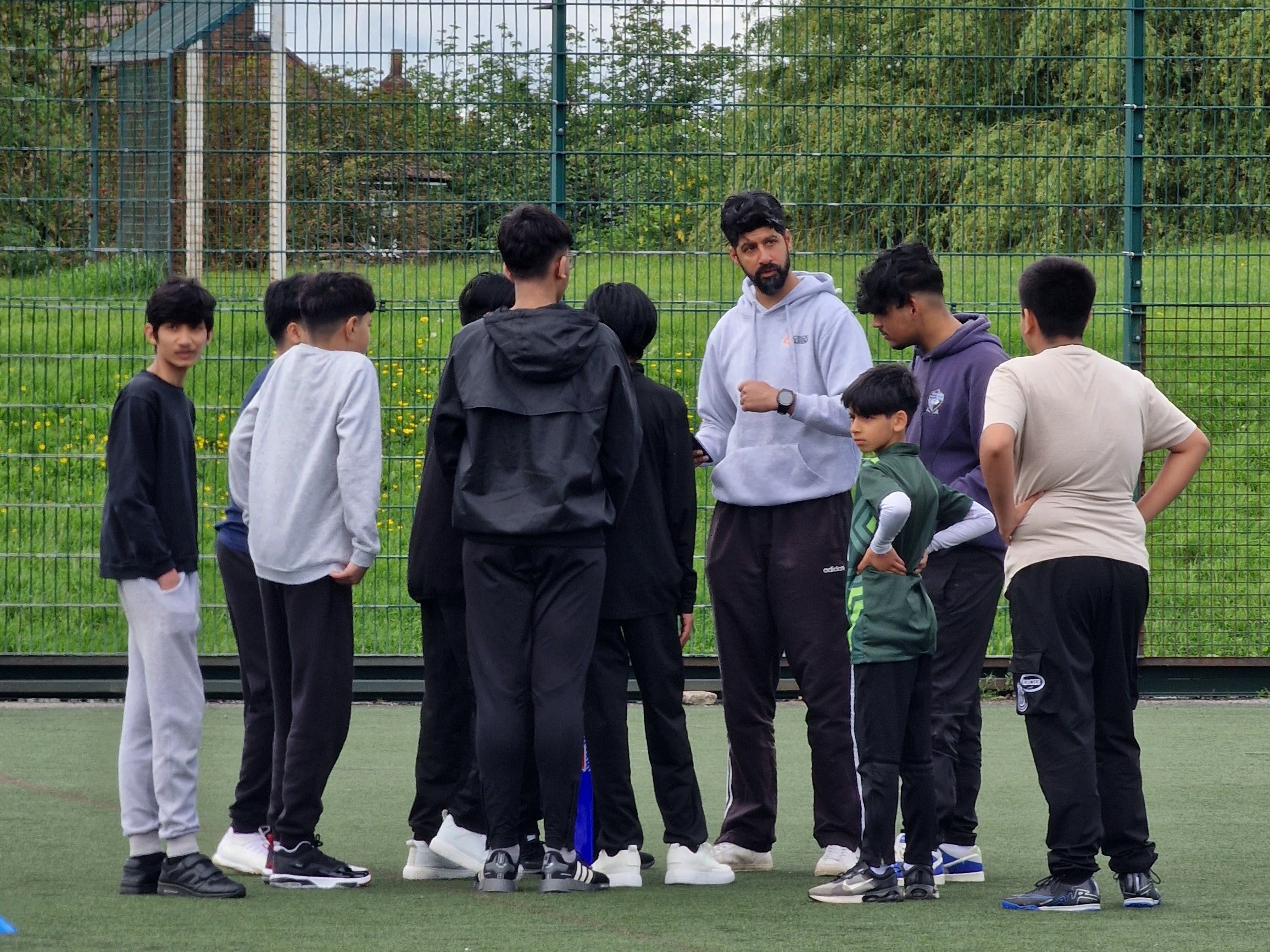 ECB coach briefs a team in a pre-match huddle, covering rules and spirit of cricket on the 3G pitch.