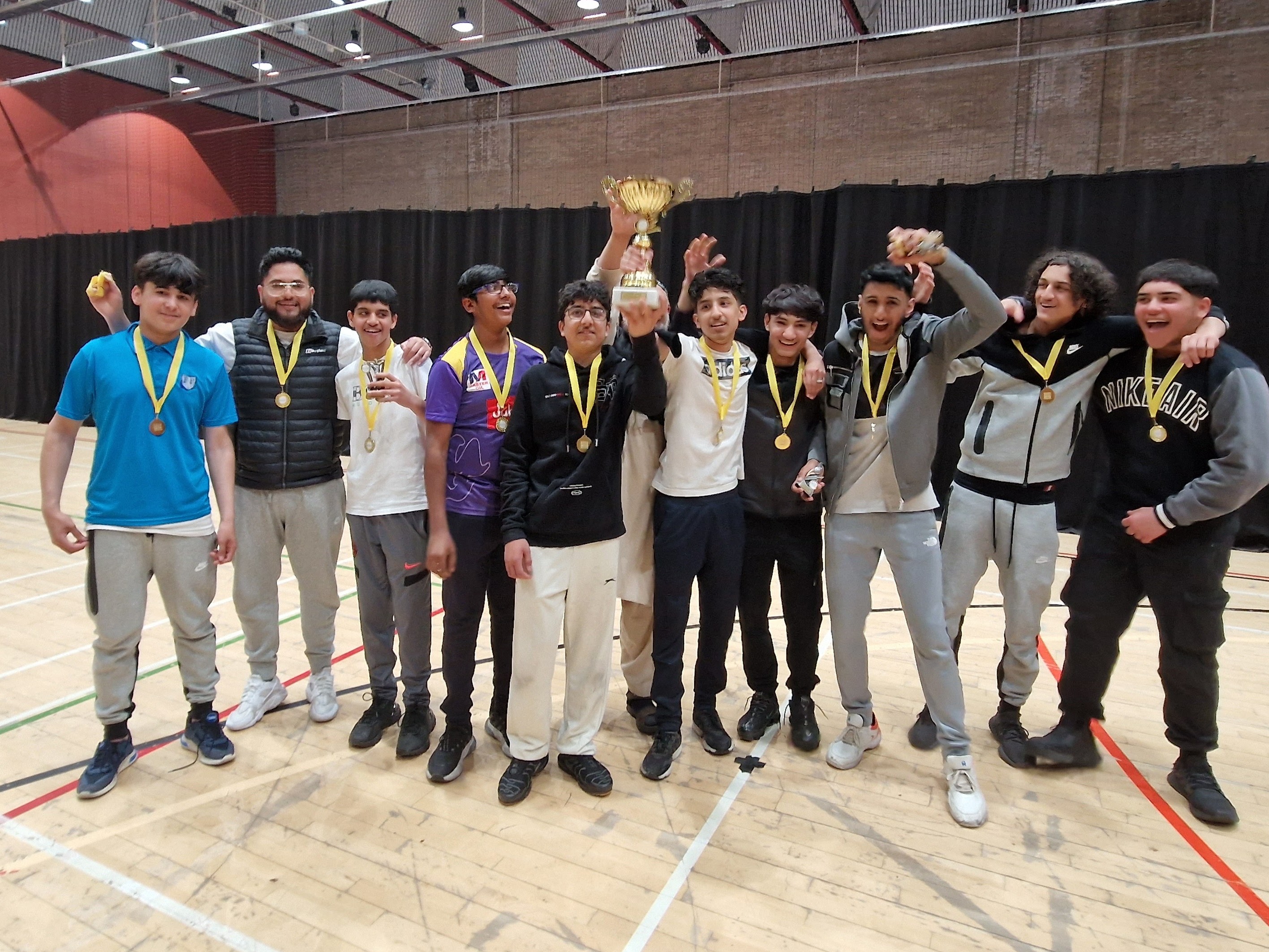 Saalik Youth celebrate indoors at Concord as the captain raises the Junior Hundred trophy; gold medals on.