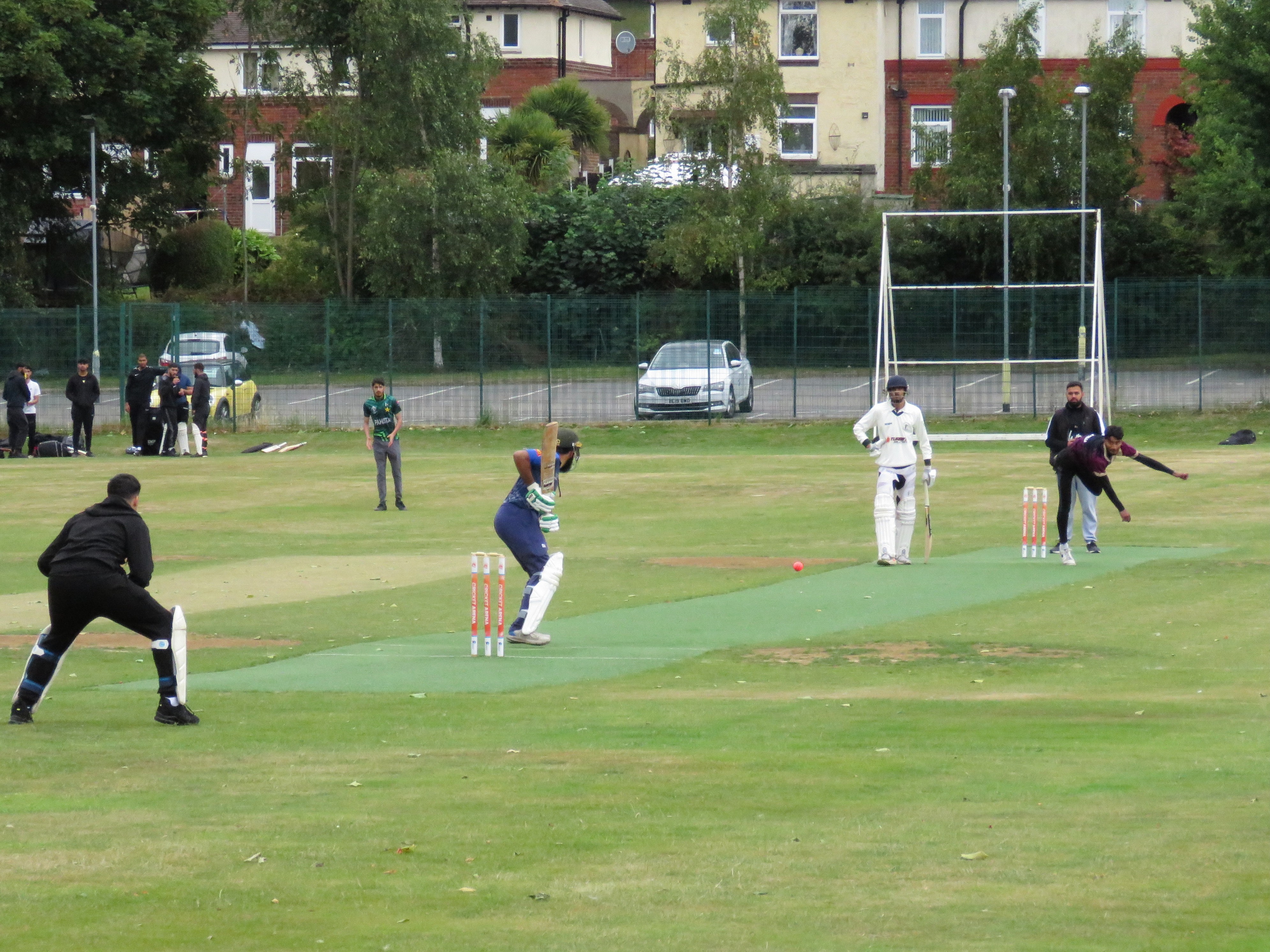 Wide shot at Shiregreen CC: bowler delivers pink hardball as batter defends on non-turf wicket during Youth Hundred in Sheffield.
