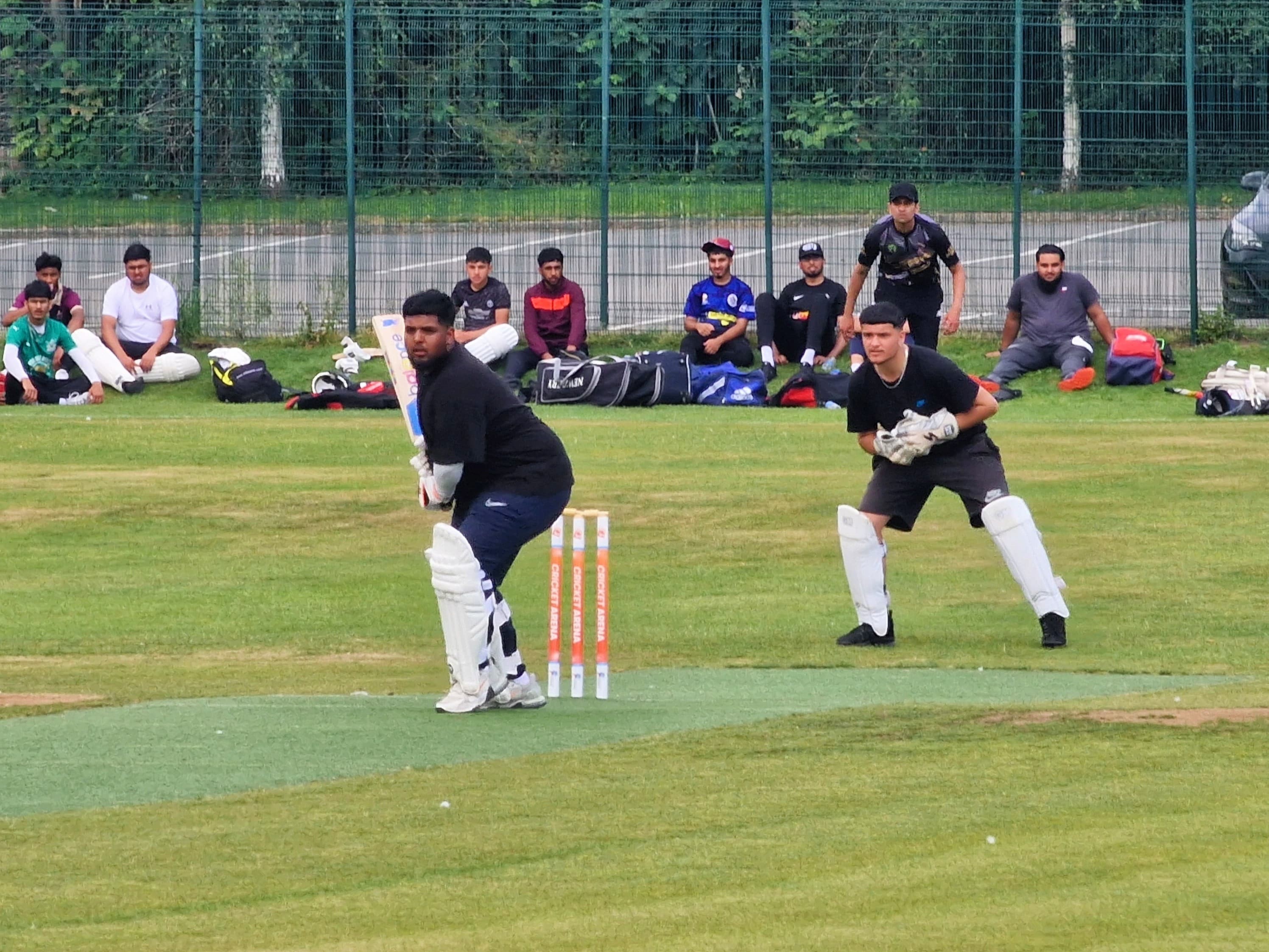 Batter and wicketkeeper ready at the crease during an outdoor hardball cricket match at Shiregreen Cricket Club, with teammates watching.