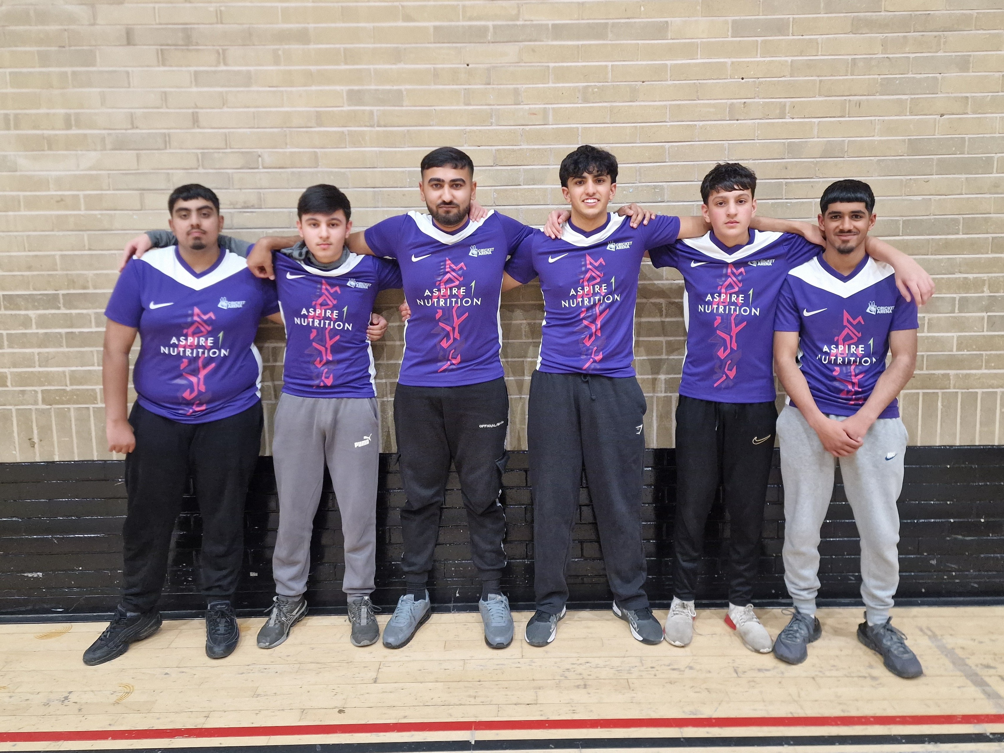 Pitsmoor Avengers youth indoor cricket team in purple Aspire 1 Nutrition Nike shirts stand arm in arm in front of a sports hall wall at Cricket Arena in Sheffield.