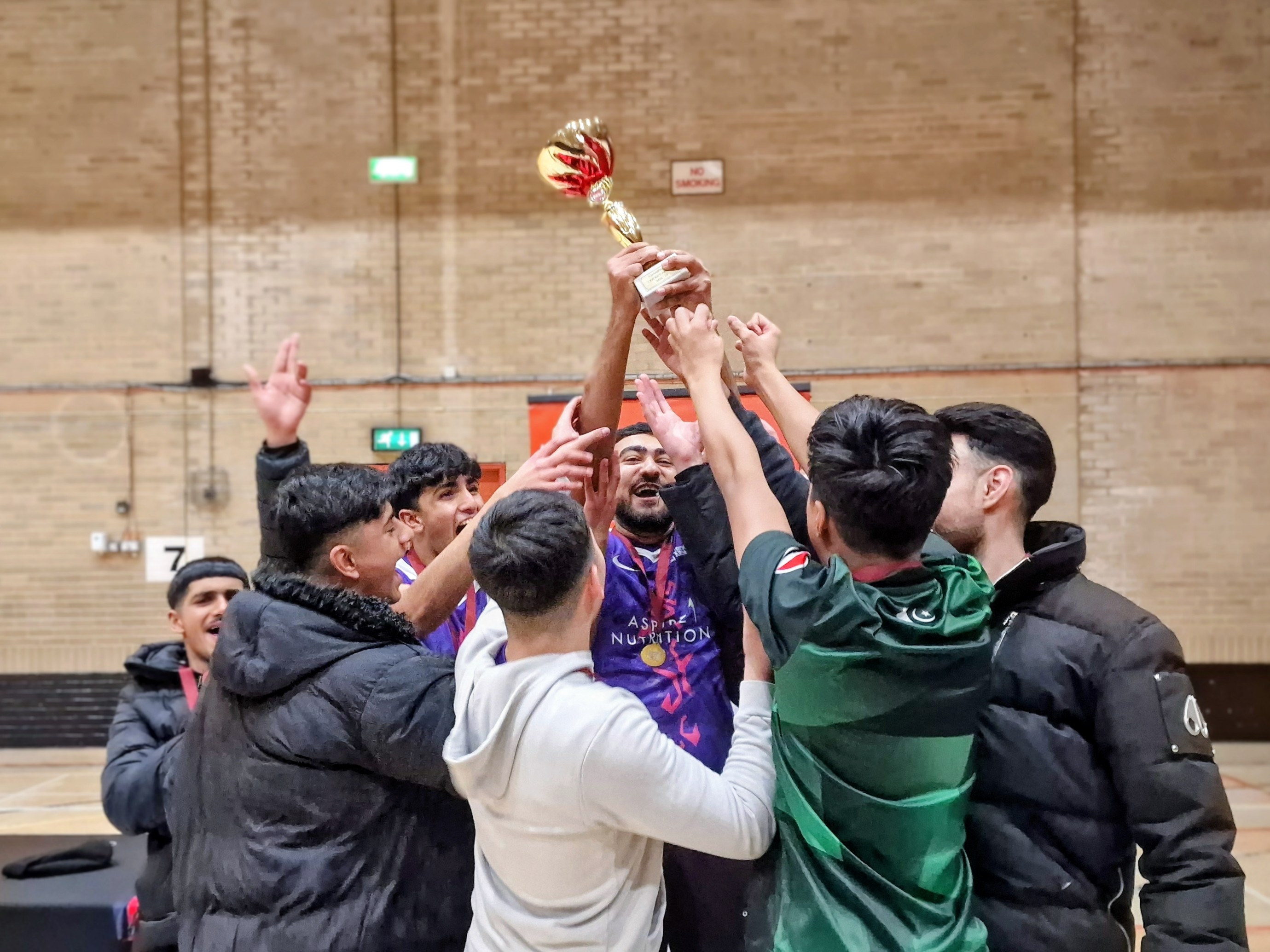 Captain Mobeen raises the MBL 2024 trophy as delighted Pitsmoor Avengers teammates reach up in celebration at Concord Sports Centre in Sheffield.