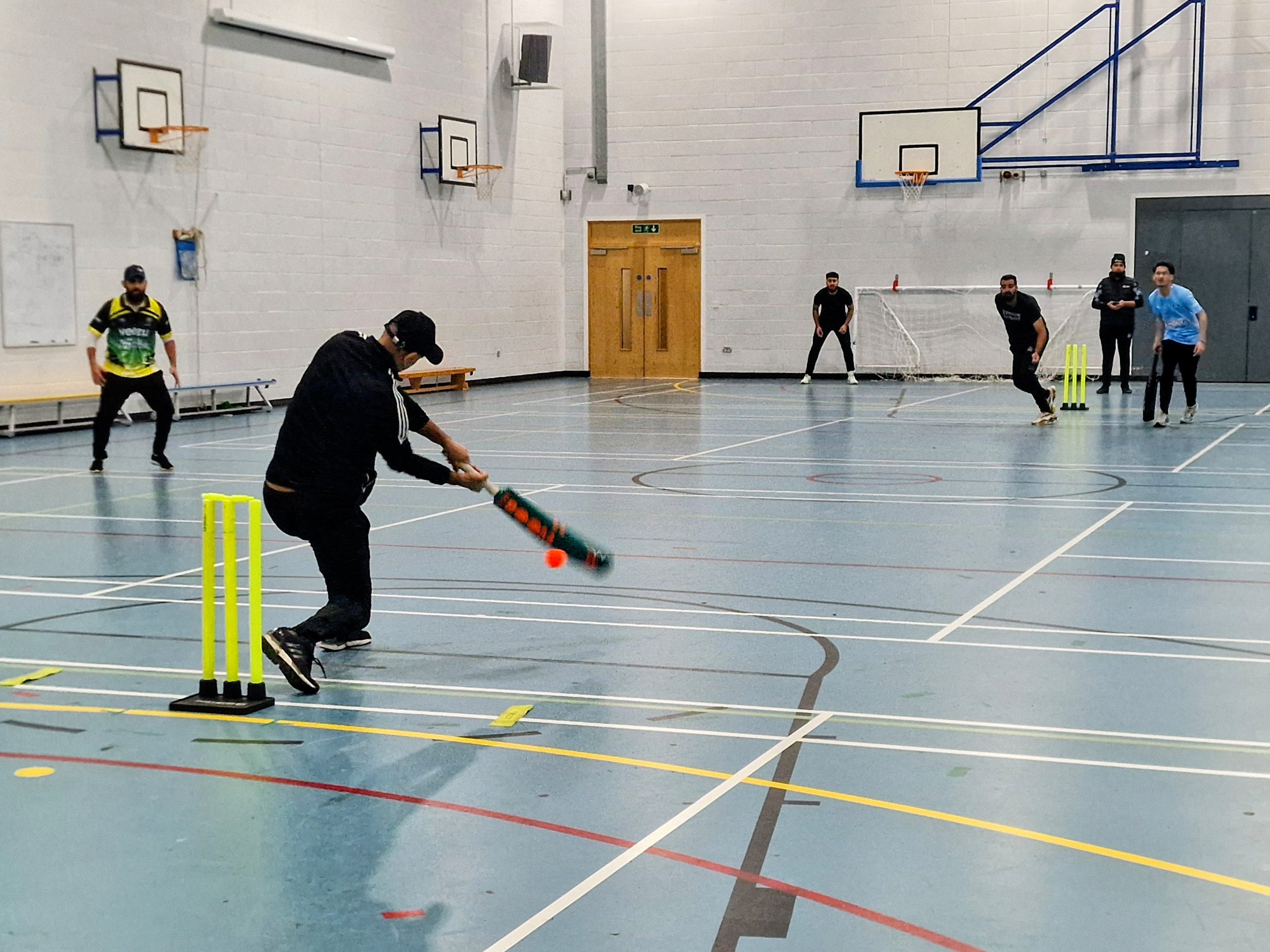 A batter plays an attacking shot in a BBICL 2024 indoor cricket match at Sheffield Park Academy, with fielders ready around the court.
